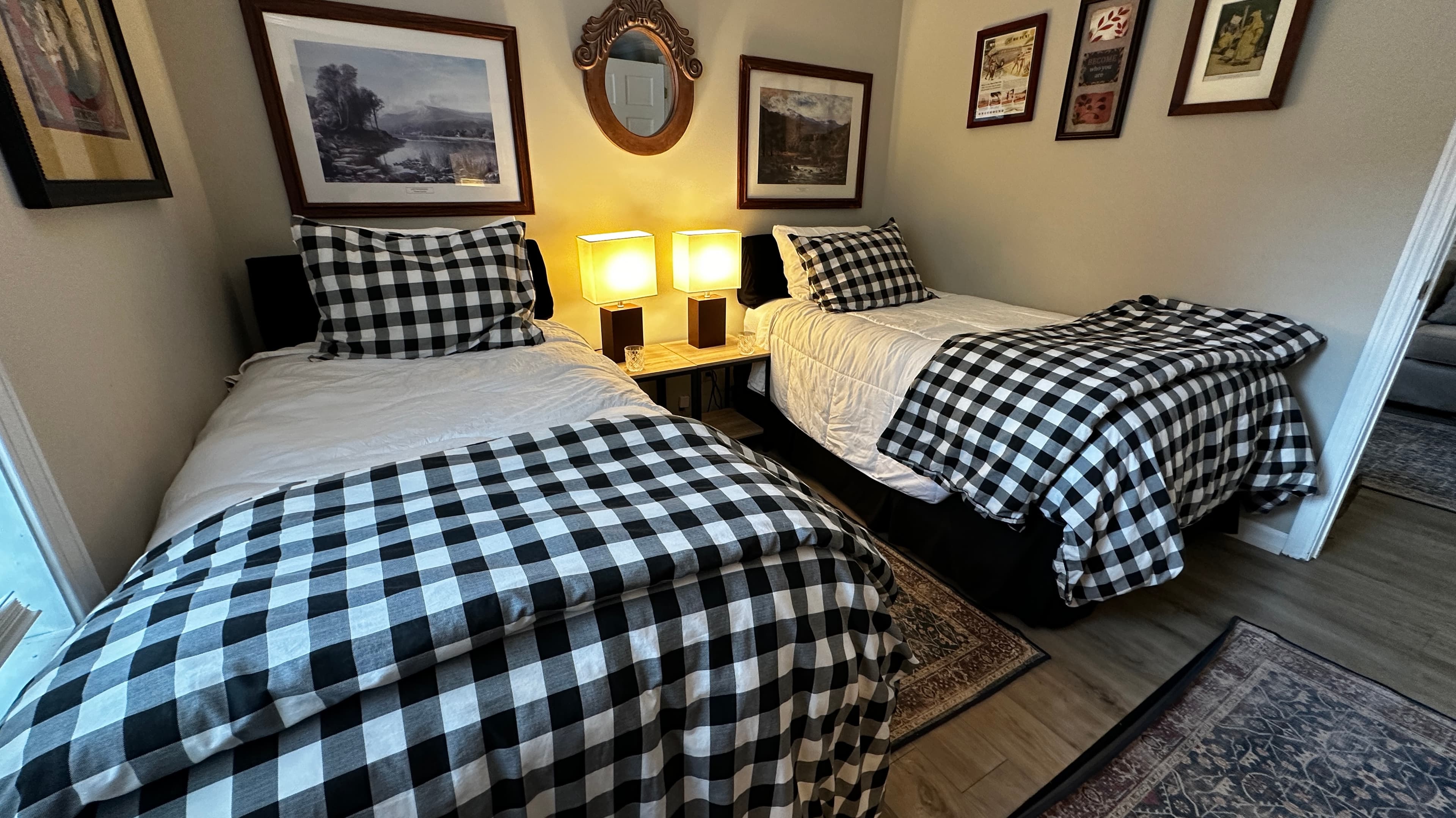 A cozy bedroom featuring two twin beds dressed in white linens and black-and-white buffalo plaid comforters. The room is decorated with multiple framed nature prints, a small oval mirror, and a central nightstand with two glowing warm lamps.