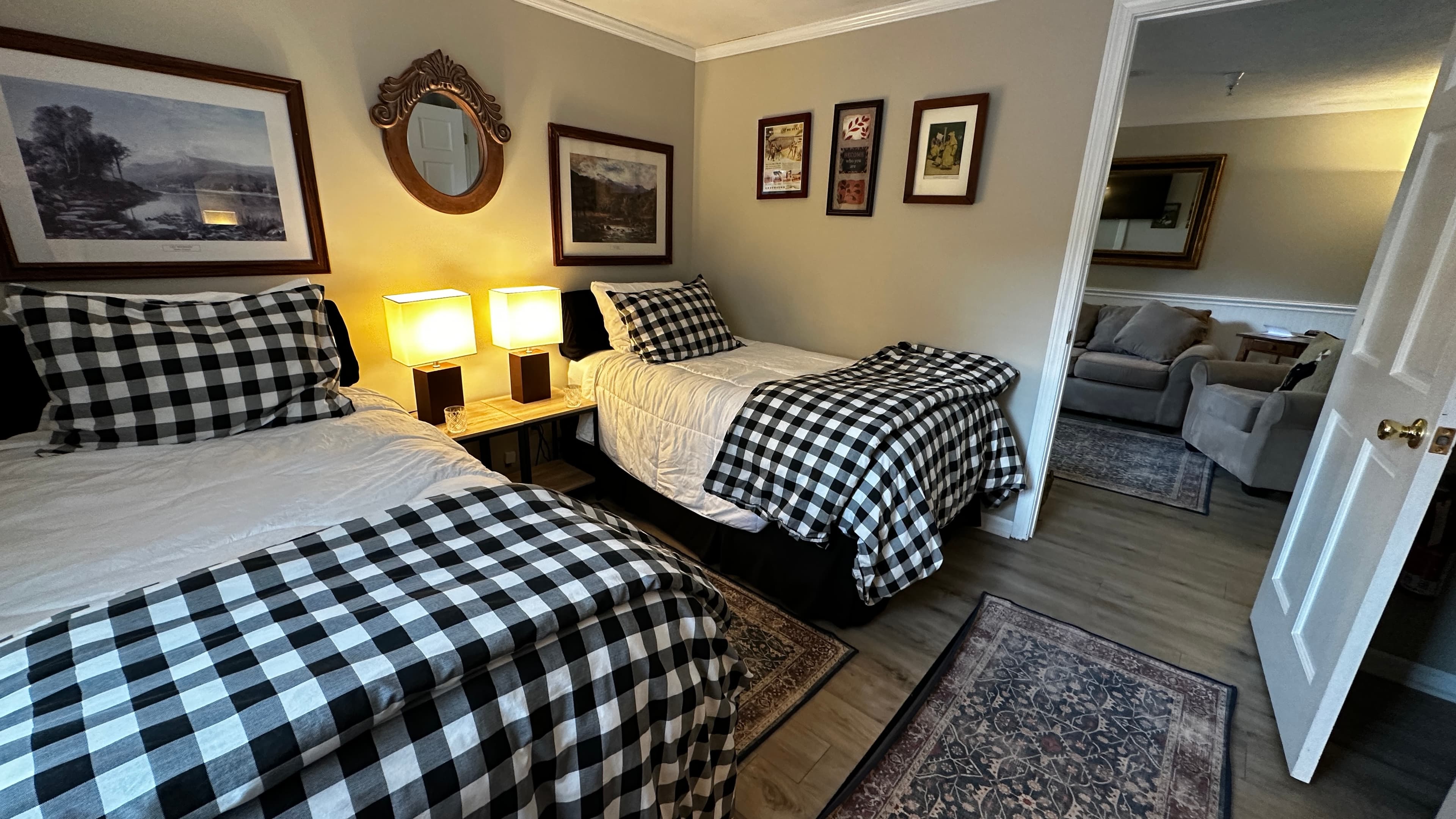 A cozy bedroom featuring two twin beds dressed in white linens and black-and-white buffalo plaid comforters. The room is decorated with multiple framed nature prints, a small oval mirror, and a central nightstand with two glowing warm lamps. An open doorway leads to a sitting area.