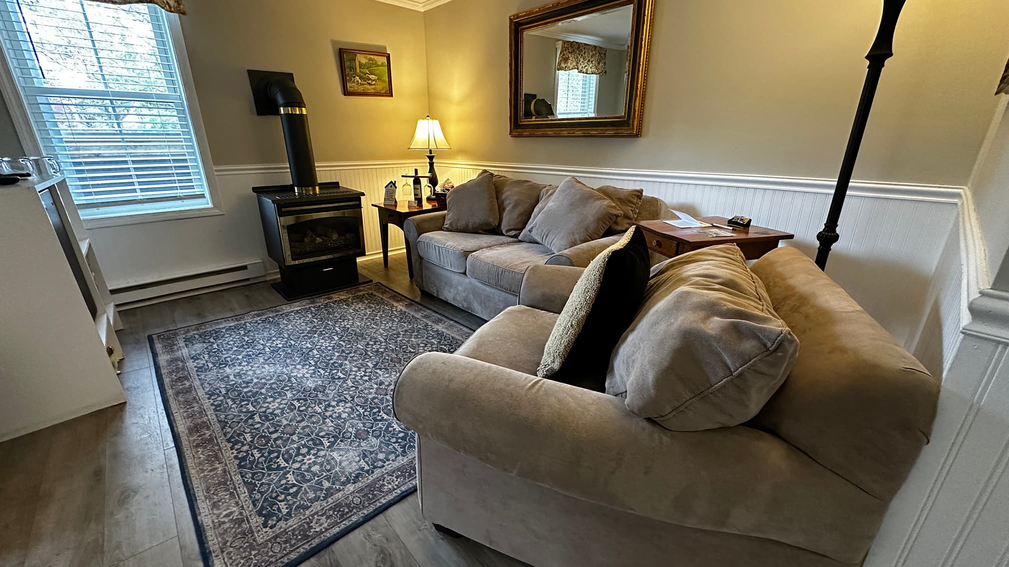 A cozy living area featuring a tan sofa and matching armchair arranged on a blue patterned area rug. The room includes white wainscoting, a gold-framed mirror, a black wood-burning stove, and a side table with a lamp casting a warm glow.