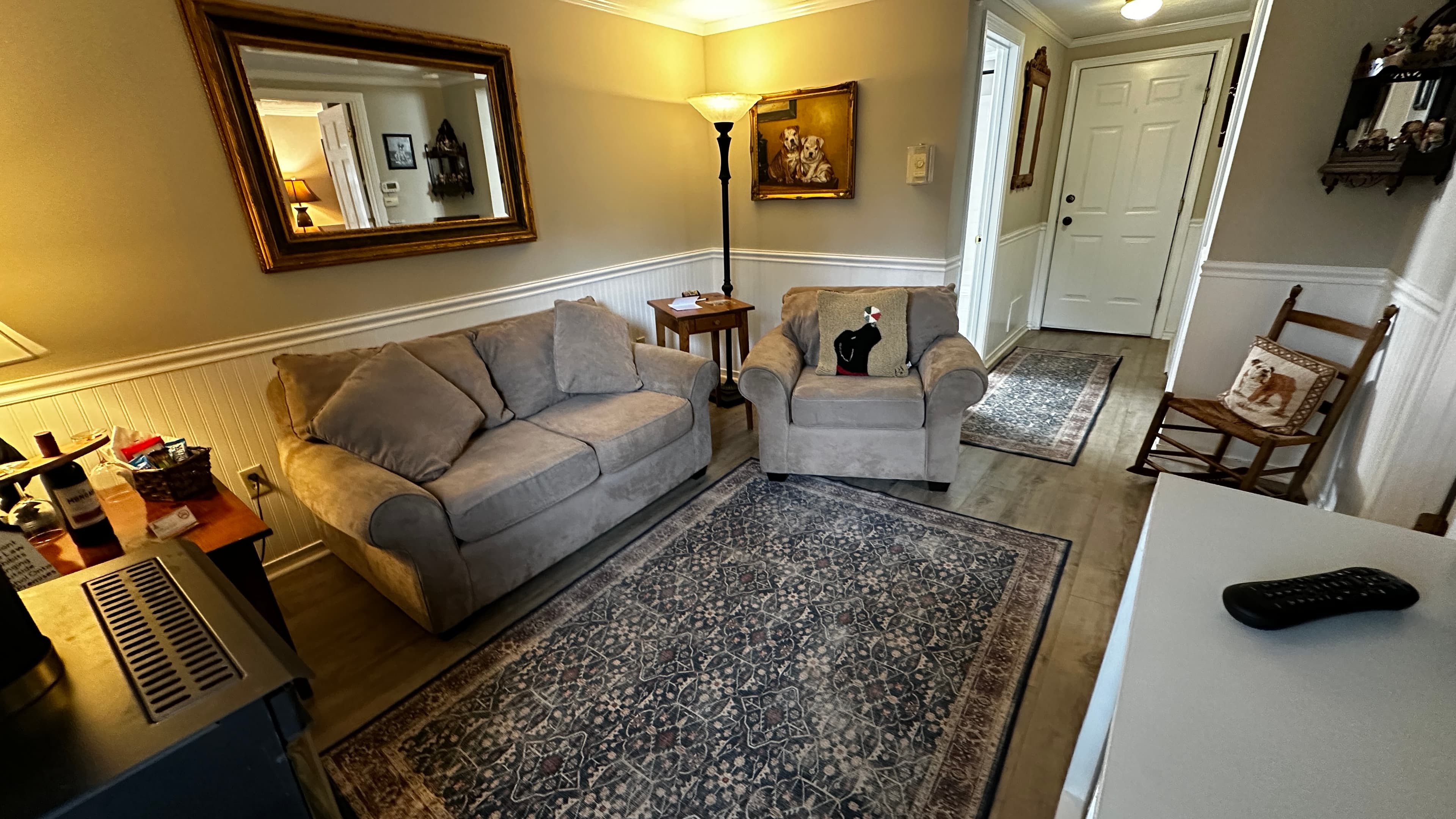 A cozy living area featuring a light grey loveseat and a matching armchair positioned on a large, intricately patterned area rug. The room includes white wainscoting, a gold-framed mirror, and a wooden rocking chair near a white front door in the background.