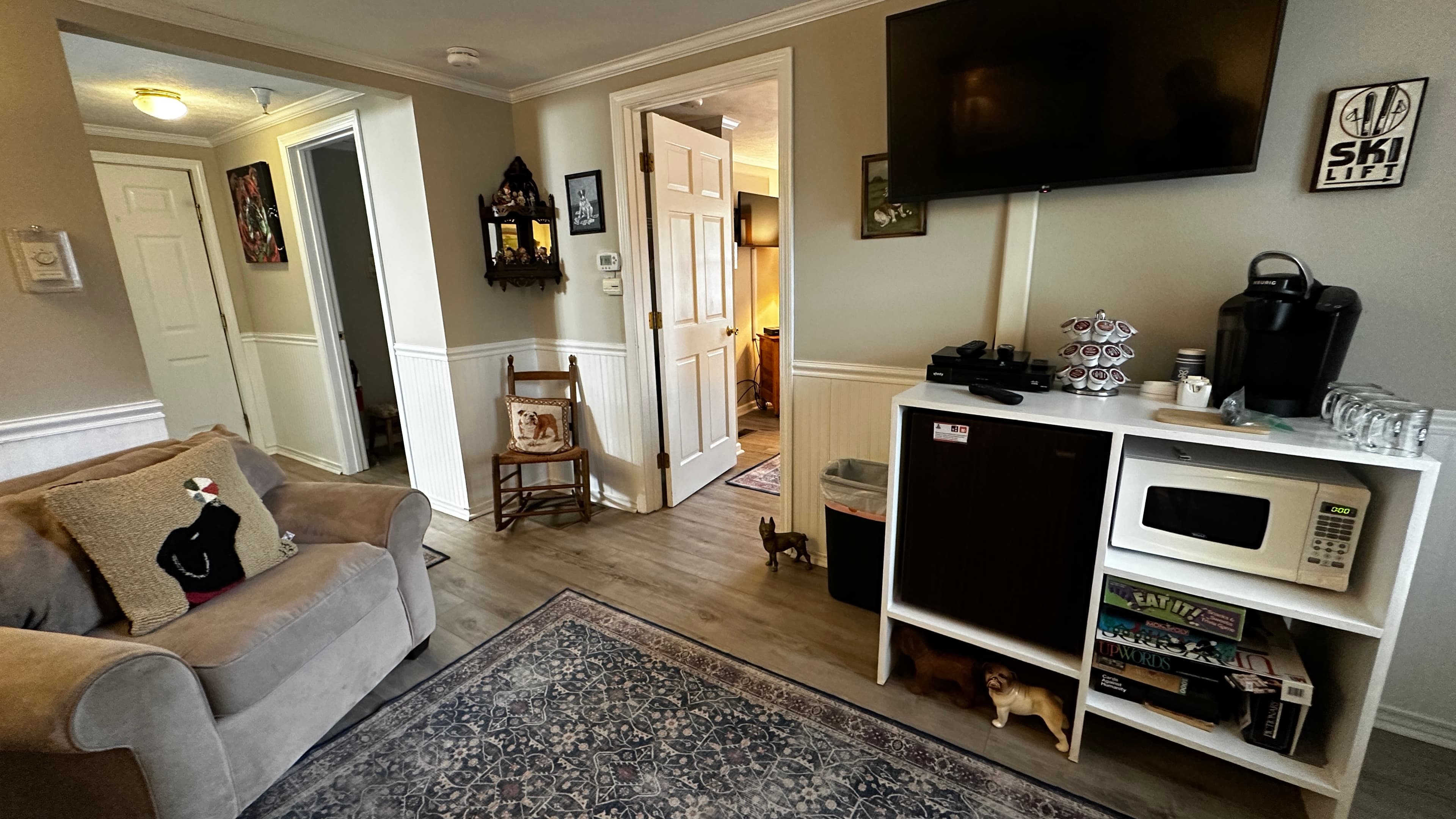 A living area featuring a light grey armchair with a black bear decorative pillow. The space includes a white storage unit equipped with a microwave, mini-fridge, and coffee maker, situated beneath a wall-mounted TV. In the background, doorways lead to other rooms and a hallway with a wooden rocking chair.
