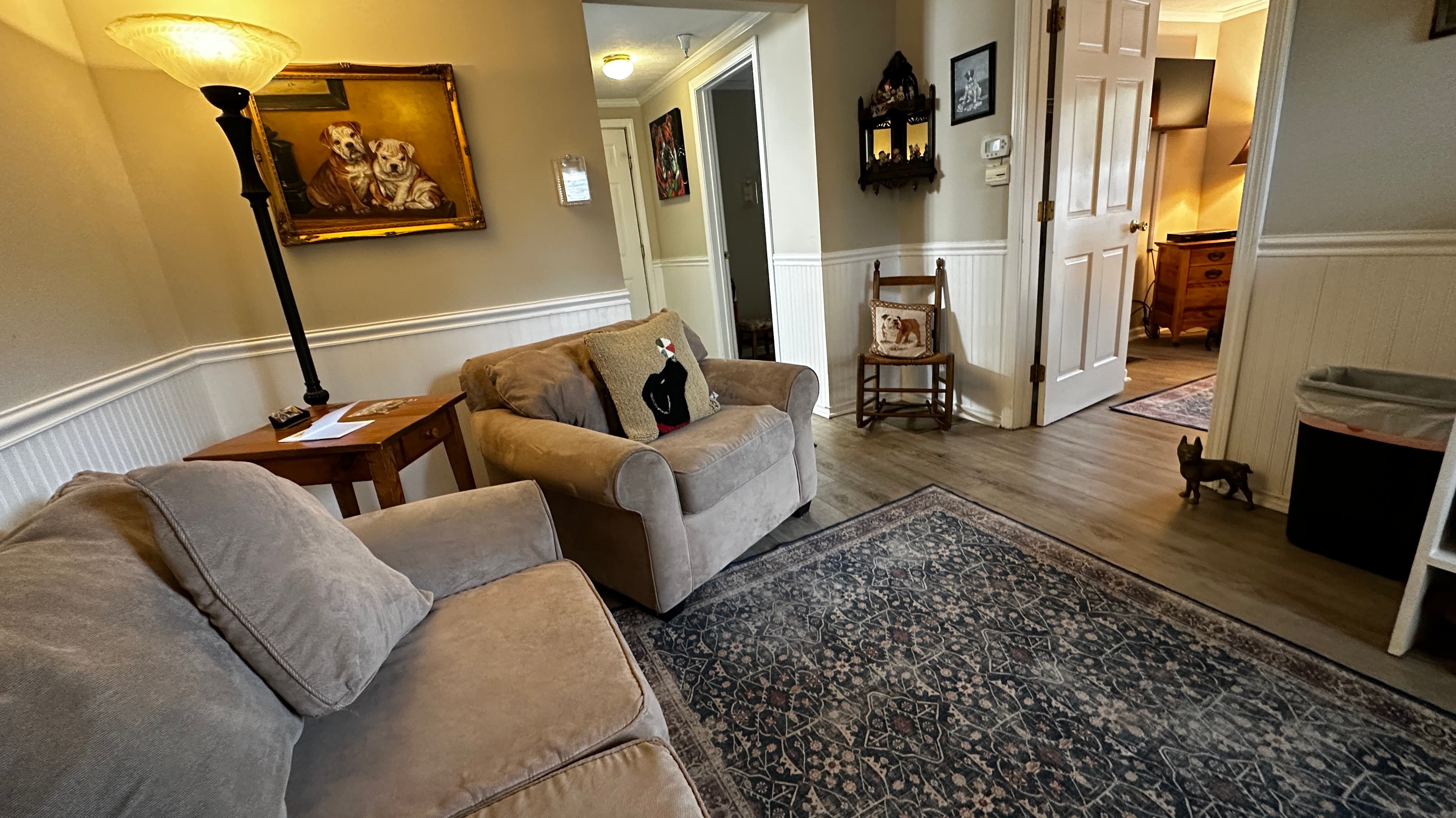 A living area featuring a light grey armchair with a black bear decorative pillow. The space includes a white storage unit equipped with a microwave, mini-fridge, and coffee maker, situated beneath a wall-mounted TV. In the background, doorways lead to other rooms and a hallway with a wooden rocking chair.
