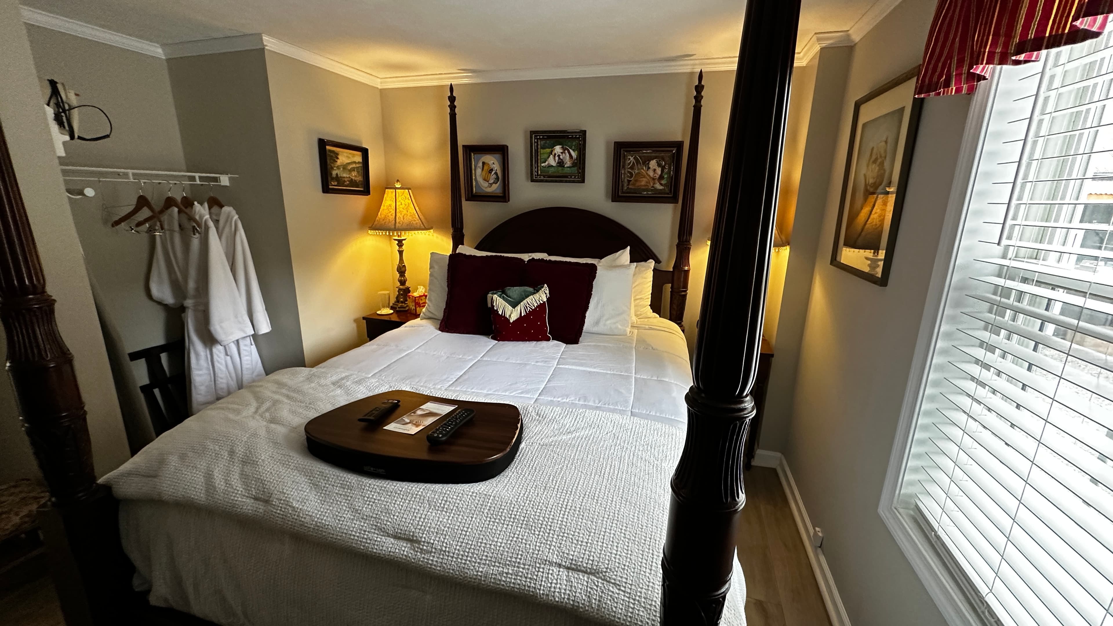 A warm and inviting bedroom with a dark wood four-poster bed made with white linens and dark red accent pillows. The space includes a clothing rack with white robes, framed artwork on the walls, and a large window with white blinds.