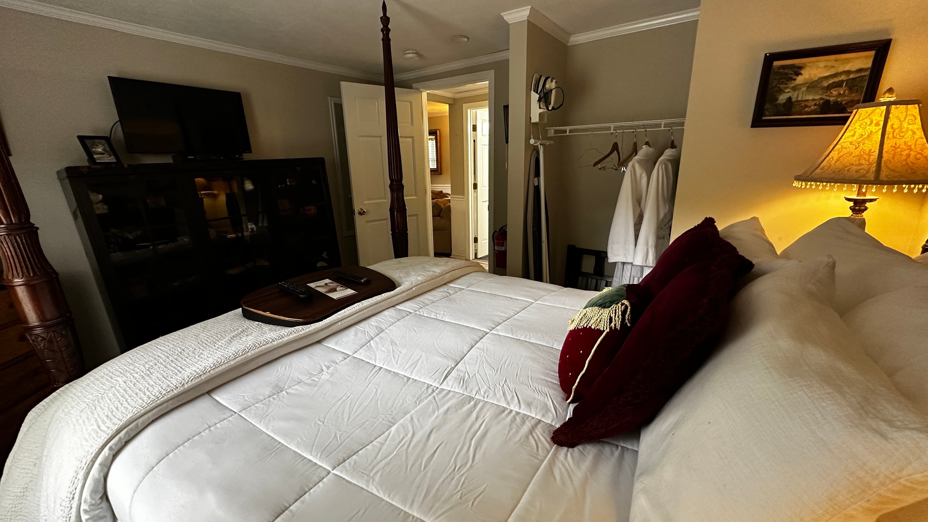 A cozy bedroom view from the head of the bed, featuring white quilted bedding and deep red accent pillows. In the background, a dark wood armoire sits beneath a wall-mounted TV, and an open doorway leads to a bright bathroom. To the right, a warm lamp illuminates a bedside area next to a clothing rack with white robes.