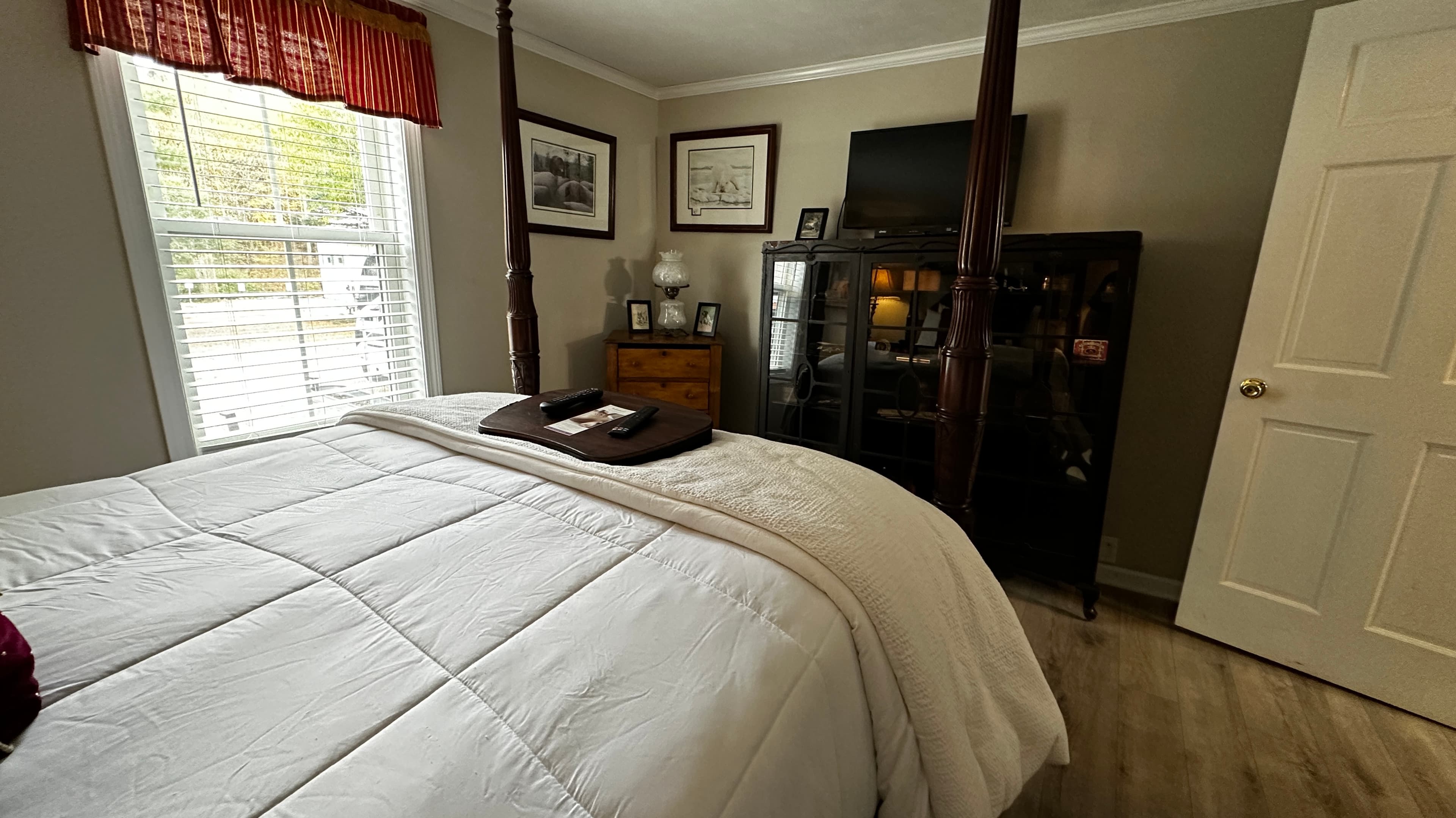 A wide-angle bedroom view from the side of a four-poster bed with white linens and a dark red accent pillow. The room features a window with a red valance, several framed pictures on the wall, and a large black armoire with a wall-mounted TV above it.