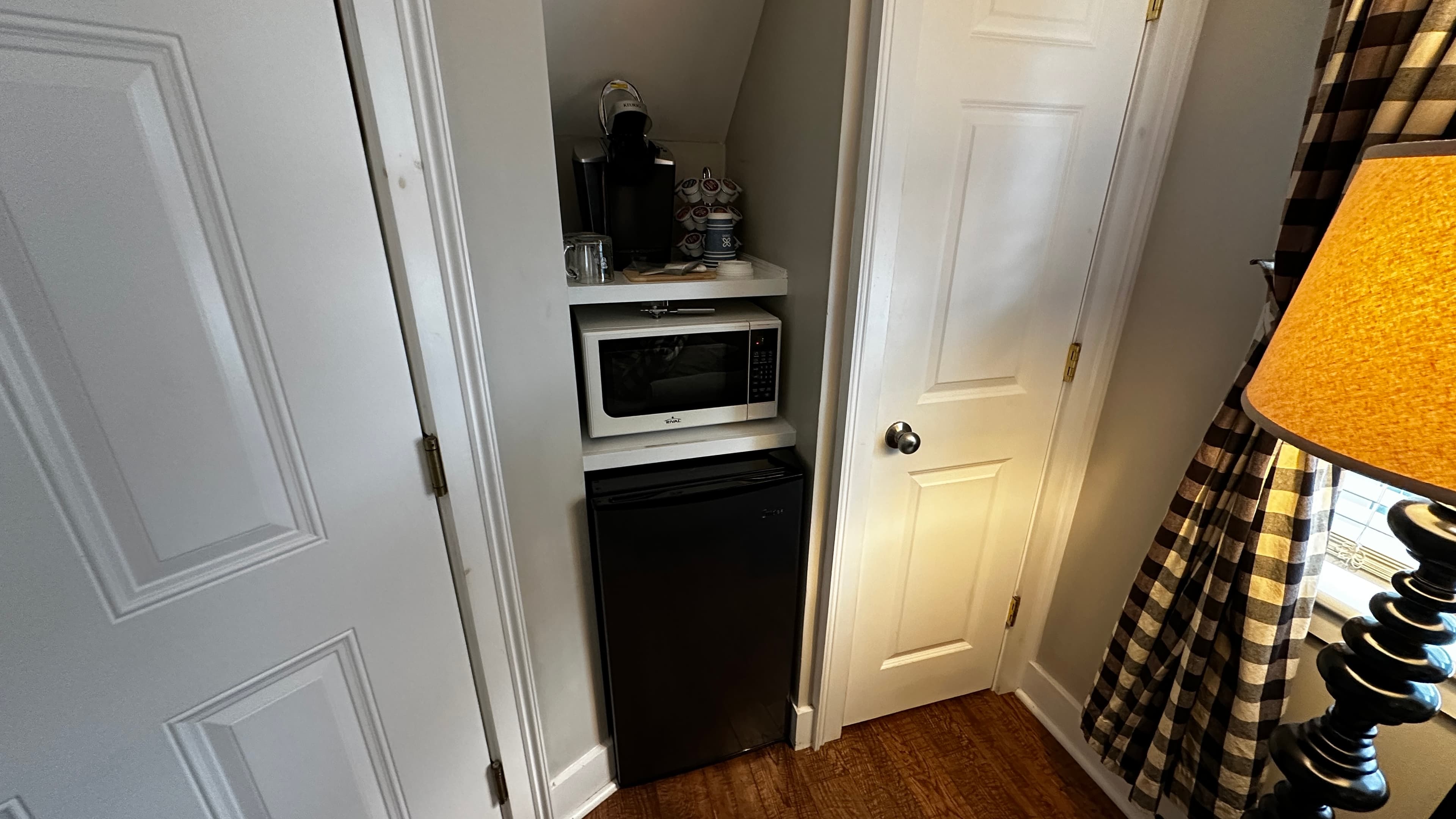 A compact kitchenette area is tucked into a wall nook between two white doors, featuring a small black refrigerator, a silver microwave, and a coffee maker on a shelf above. A dark wood lamp with a checkered shade is visible in the foreground to the right.
