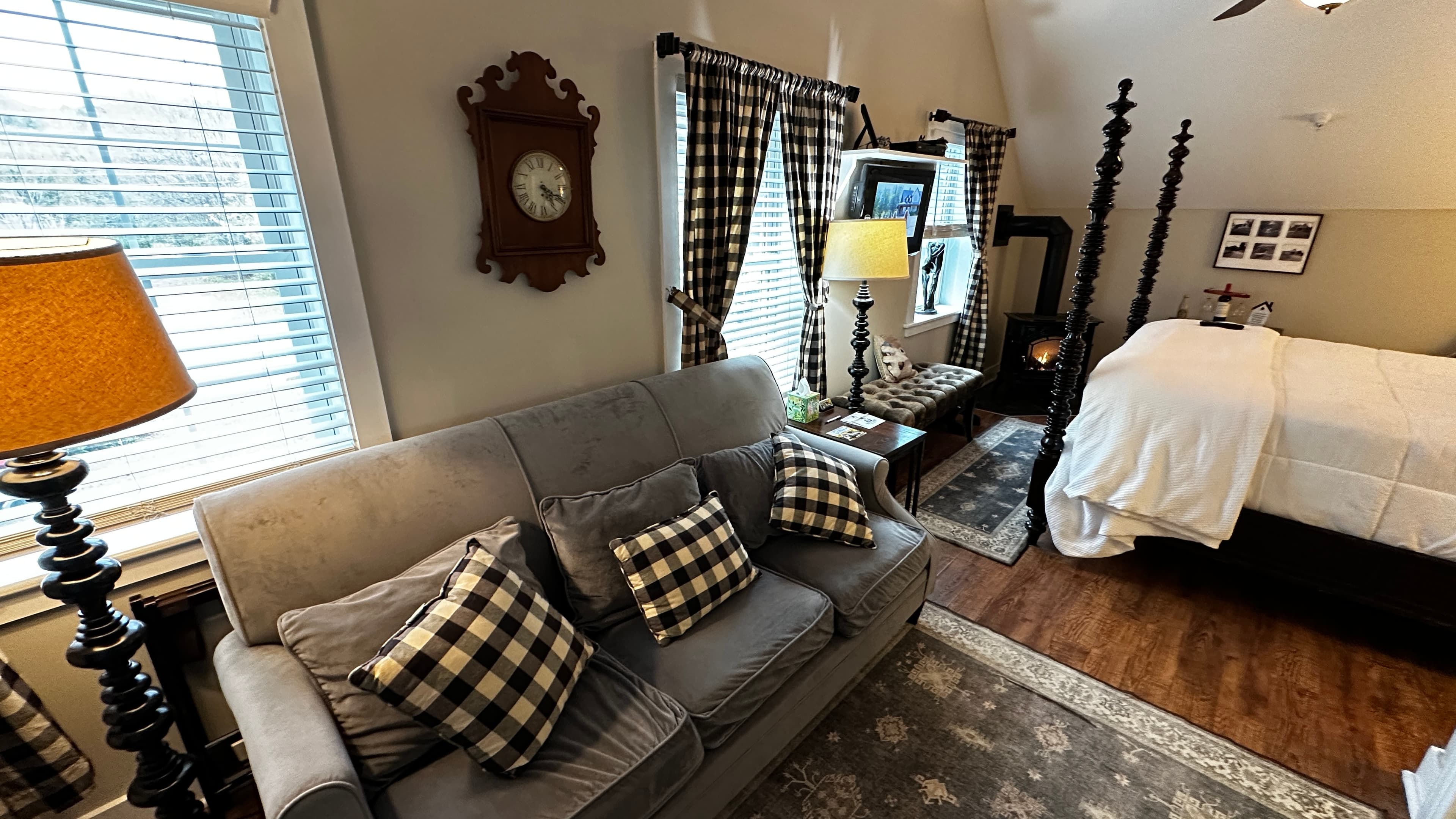A cozy seating area in an attic-style suite, featuring a plush gray sofa with black-and-white checkered pillows. The room includes a traditional wall clock, windows with checkered curtains, and a view into the bedroom area featuring a dark wood four-poster bed.