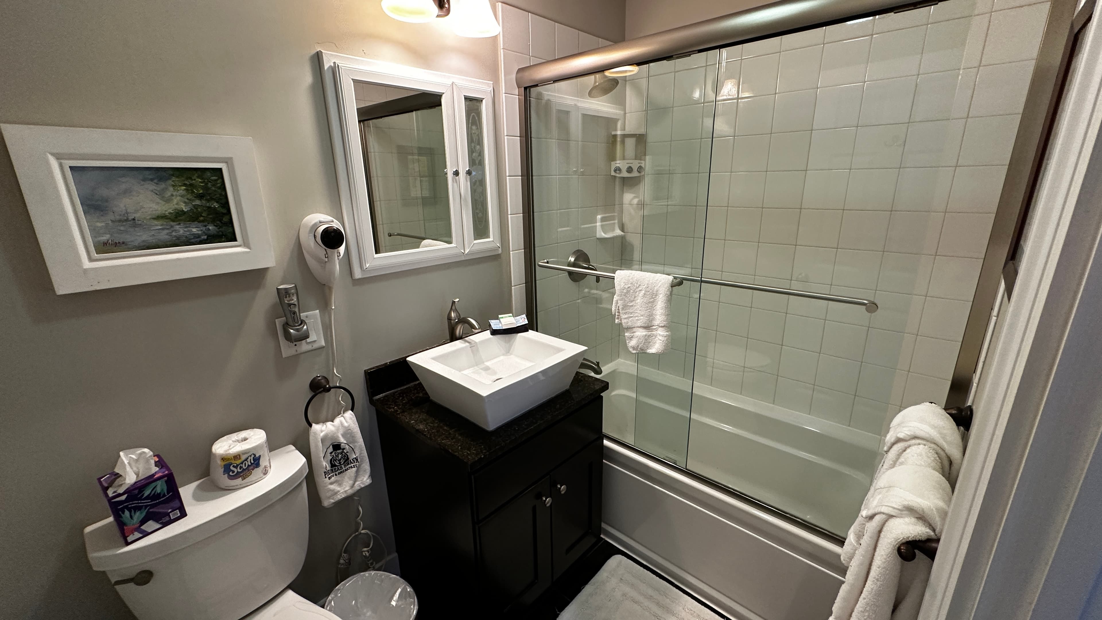 A bathroom featuring a modern square vessel sink on a dark wood vanity and a bathtub-shower combination with sliding glass doors. The room includes a white toilet, neutral gray walls, and white towels hanging on the shower door handle.