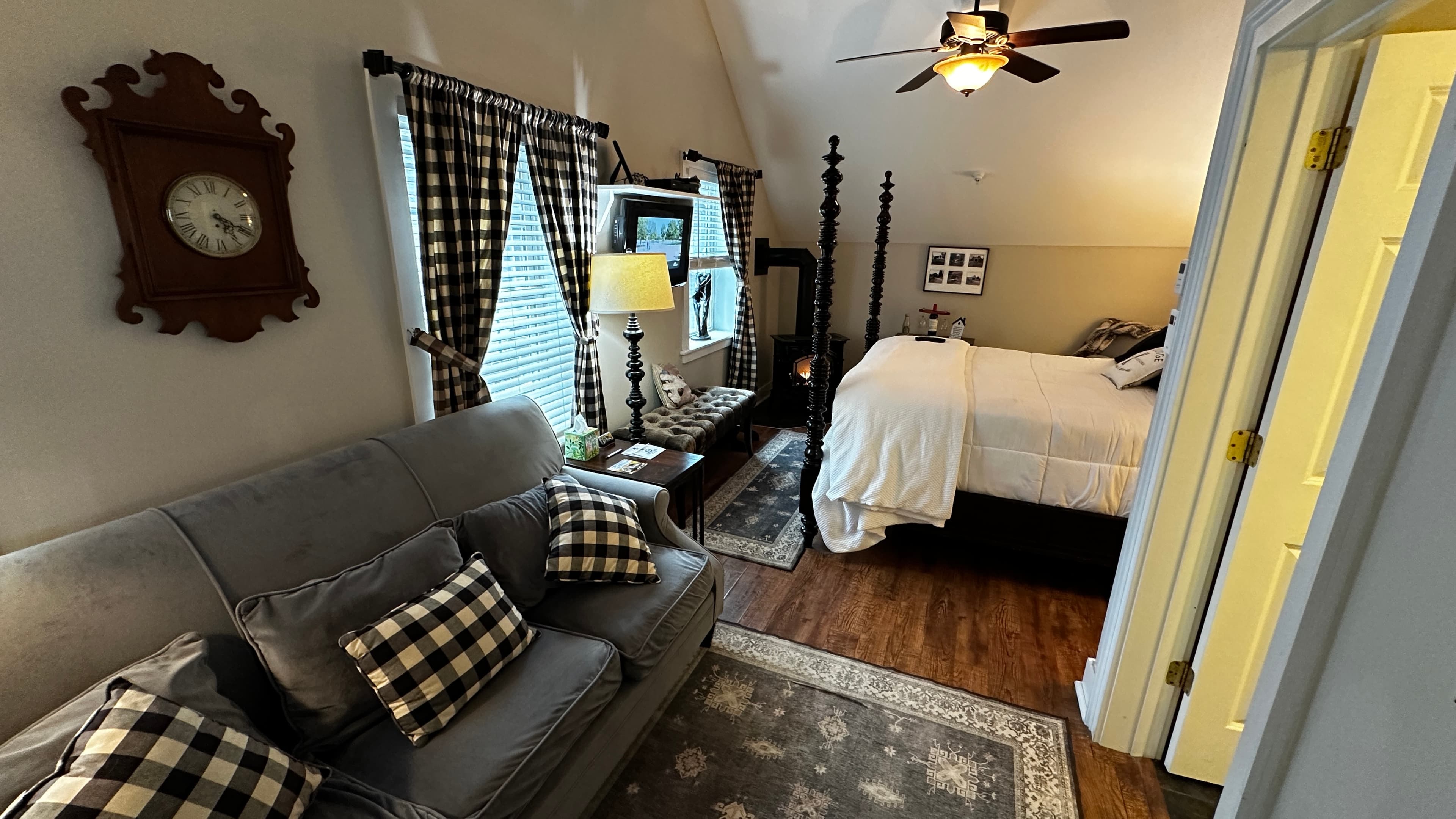 A comfortable suite featuring a gray sofa with buffalo plaid pillows, a four-poster bed with white linens, and a vaulted ceiling with a wooden ceiling fan. The room is decorated in a farmhouse style with wood flooring, plaid curtains, and a large vintage wall clock.