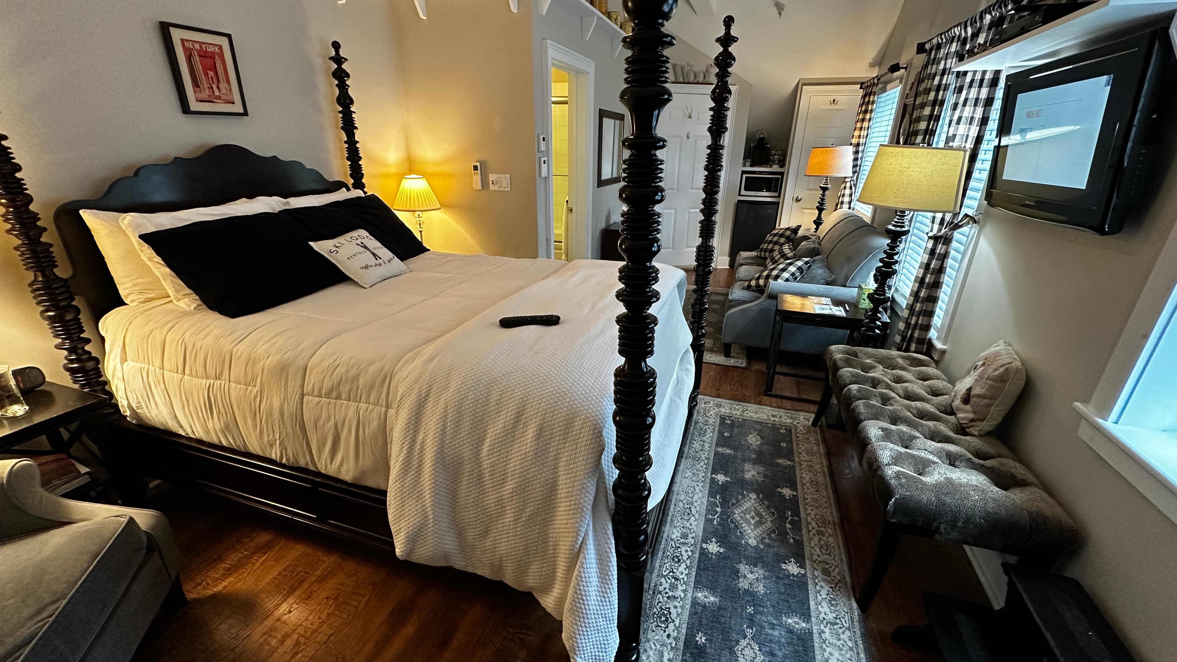 A welcoming bedroom featuring a dark wood four-poster bed with white and black bedding. The room includes a narrow patterned rug, a cushioned storage bench at the foot of the bed, and warm lighting from bedside lamps against neutral walls.