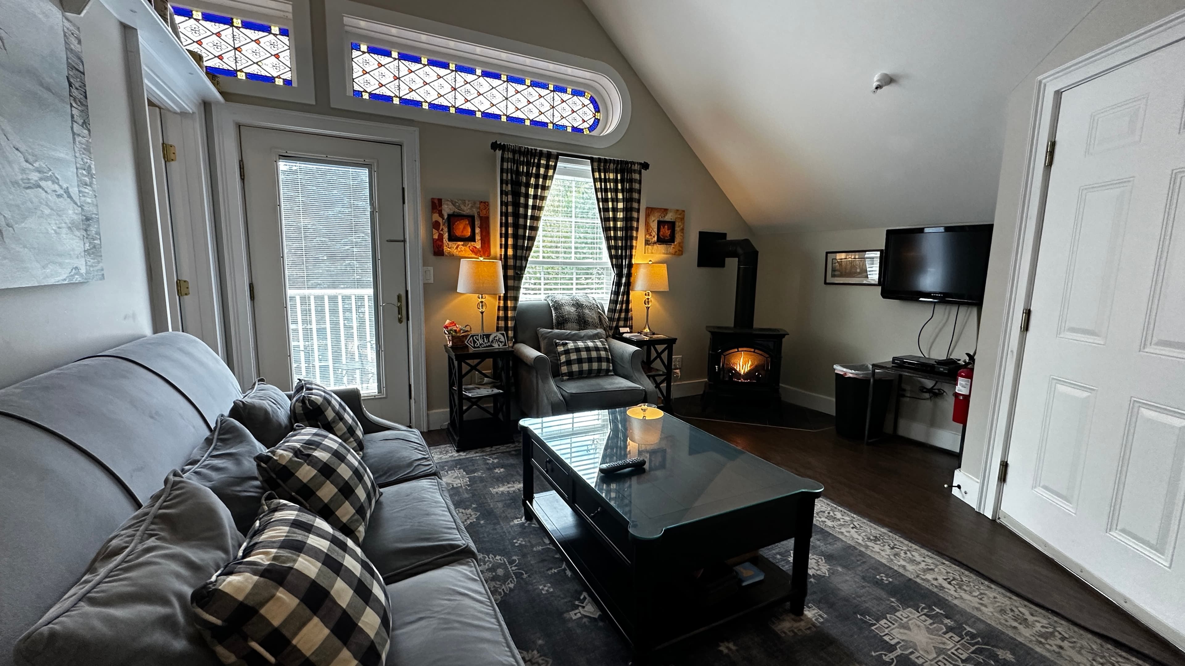 A cozy living room with a vaulted ceiling featuring a grey sofa with buffalo plaid pillows and a dark wood coffee table on a patterned rug. The room includes a black gas log stove with a visible fire, a wall-mounted TV, and unique stained-glass transom windows above a white door leading to a balcony.