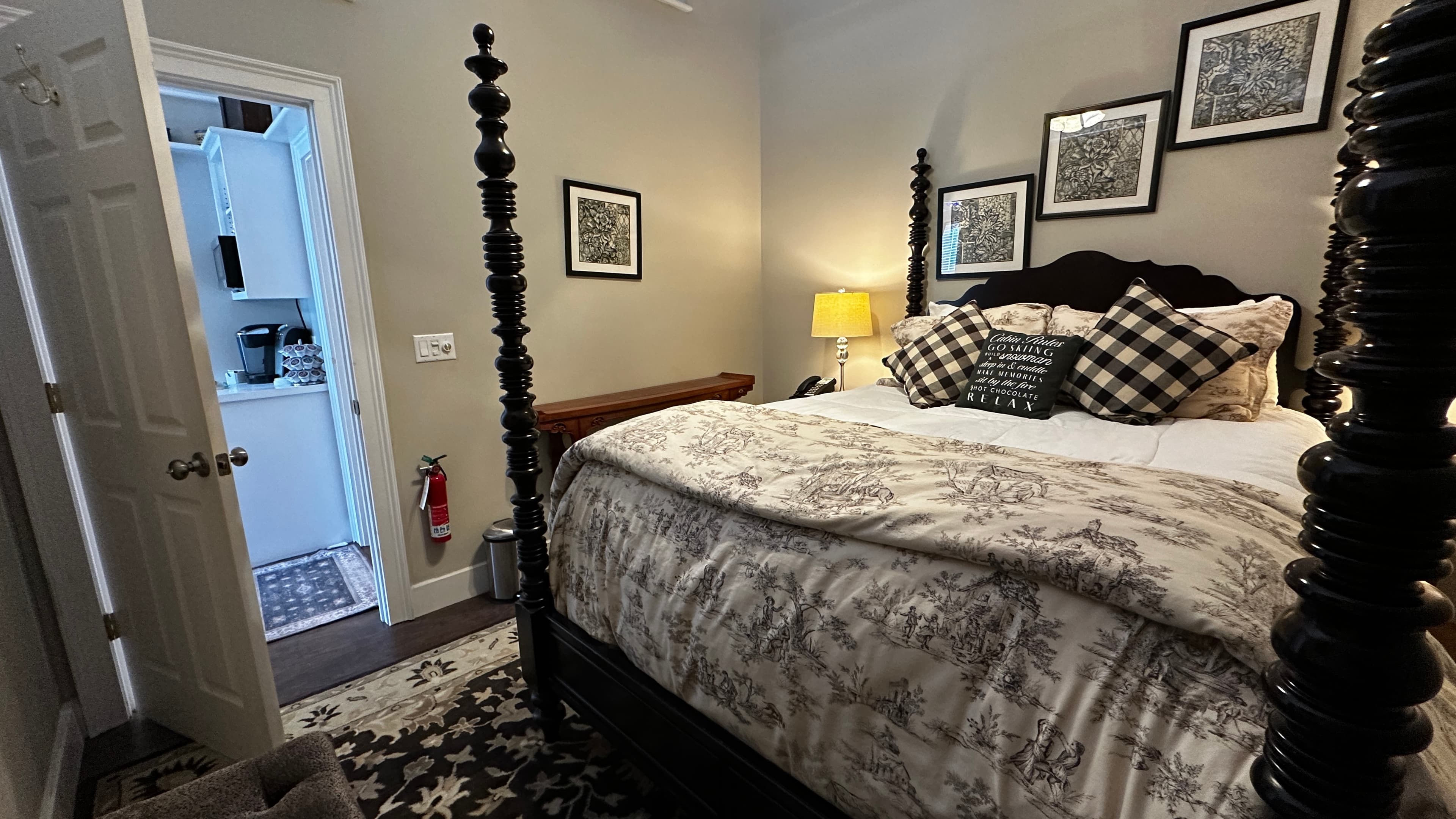 A cozy bedroom featuring a black four-poster bed with a patterned gray and white duvet and buffalo plaid accent pillows. The room includes several framed black-and-white photos on the wall, a bedside lamp casting a warm glow, and an open doorway leading to a brightly lit kitchenette area.