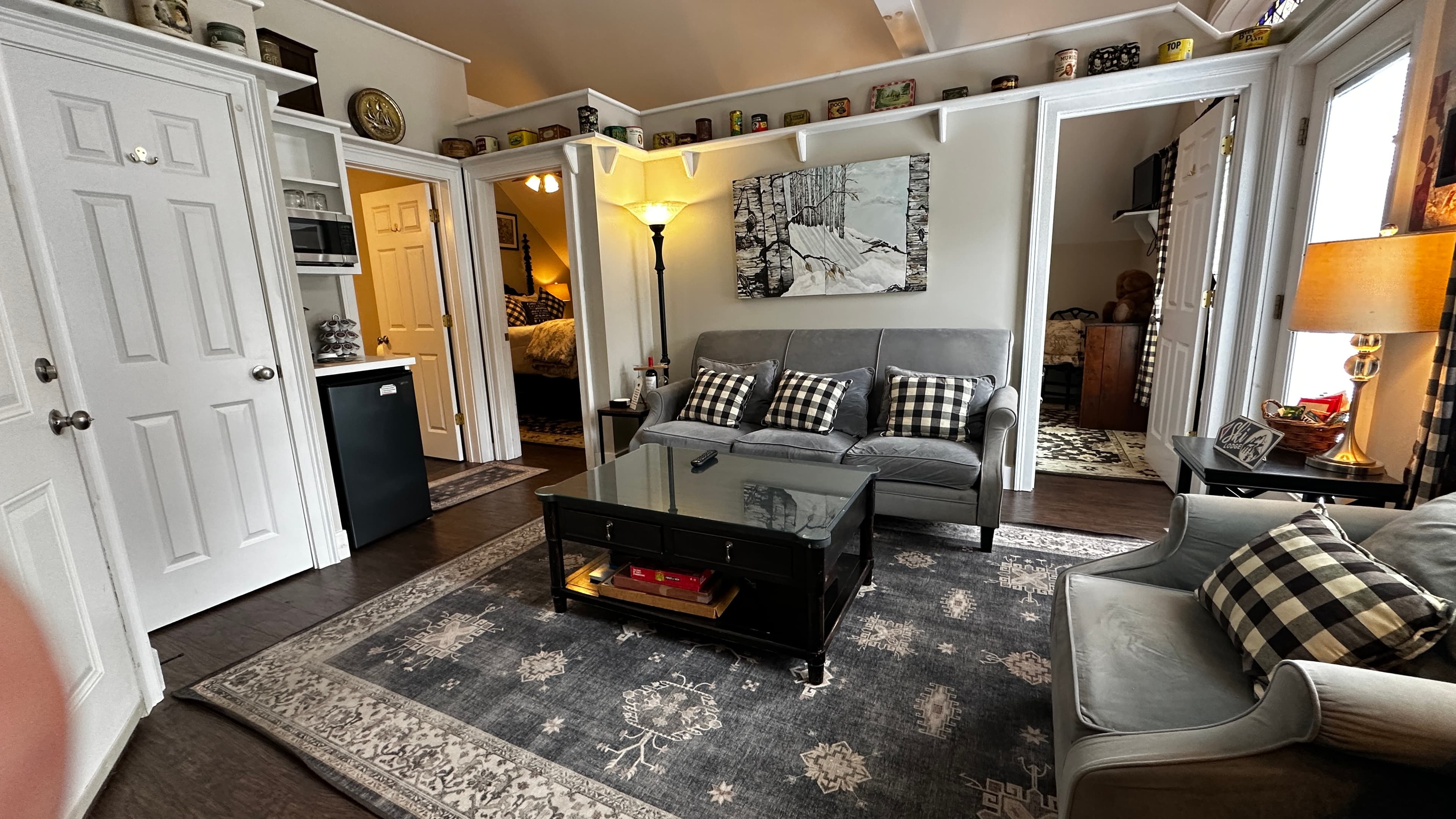 A spacious, farmhouse-style living room with a vaulted ceiling, featuring a grey sofa and armchair accented with buffalo plaid pillows. The room includes a black glass coffee table on a large patterned rug, a small kitchenette with a mini-fridge, and an open doorway leading to a bedroom.