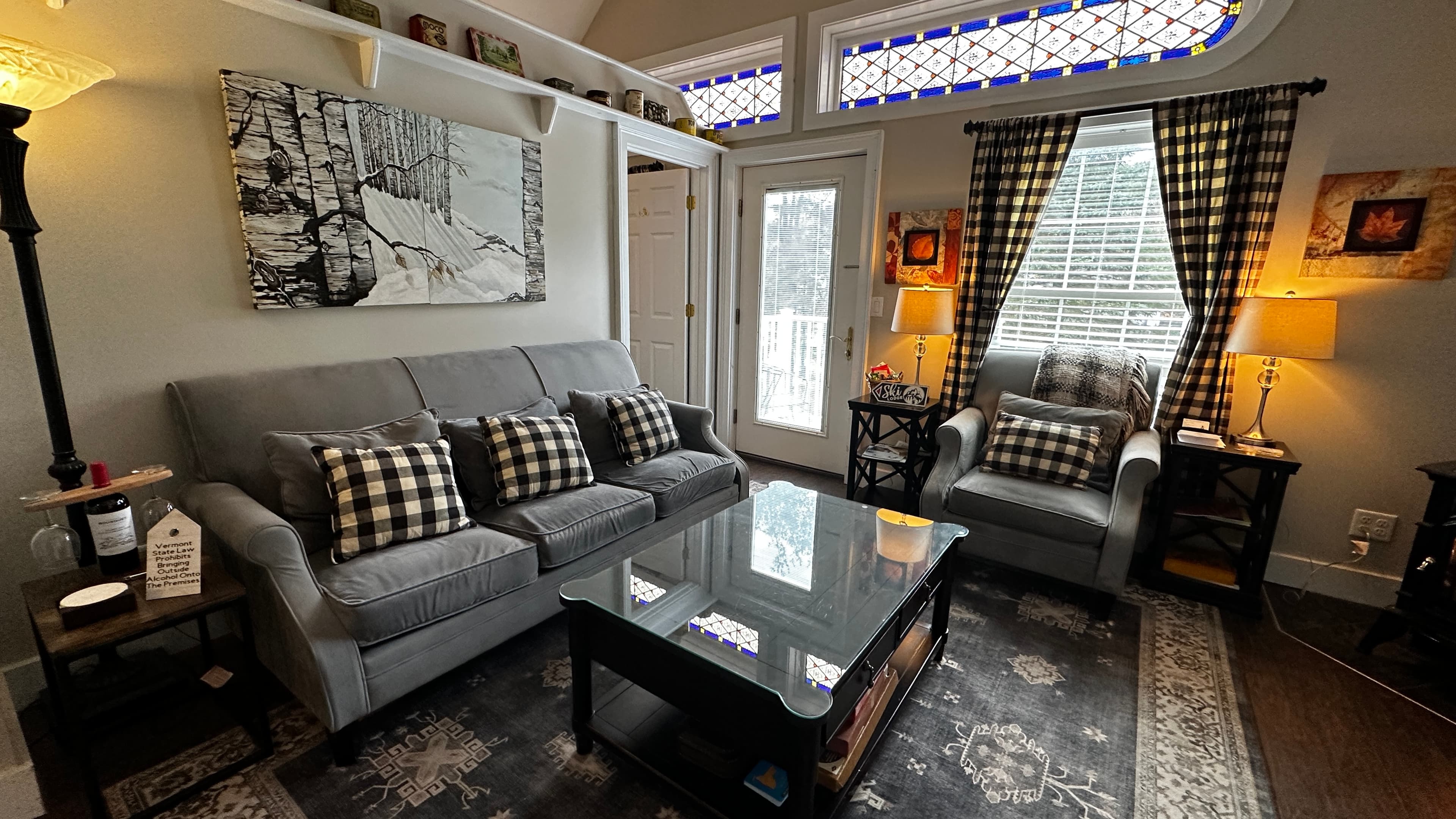 A farmhouse-style living room featuring a long gray sofa with buffalo plaid pillows and a glass-top coffee table on a large patterned area rug. The room is decorated with a large winter-themed wall art piece, warm lamps, and a unique stained-glass transom window above a white door leading to an outdoor space.
