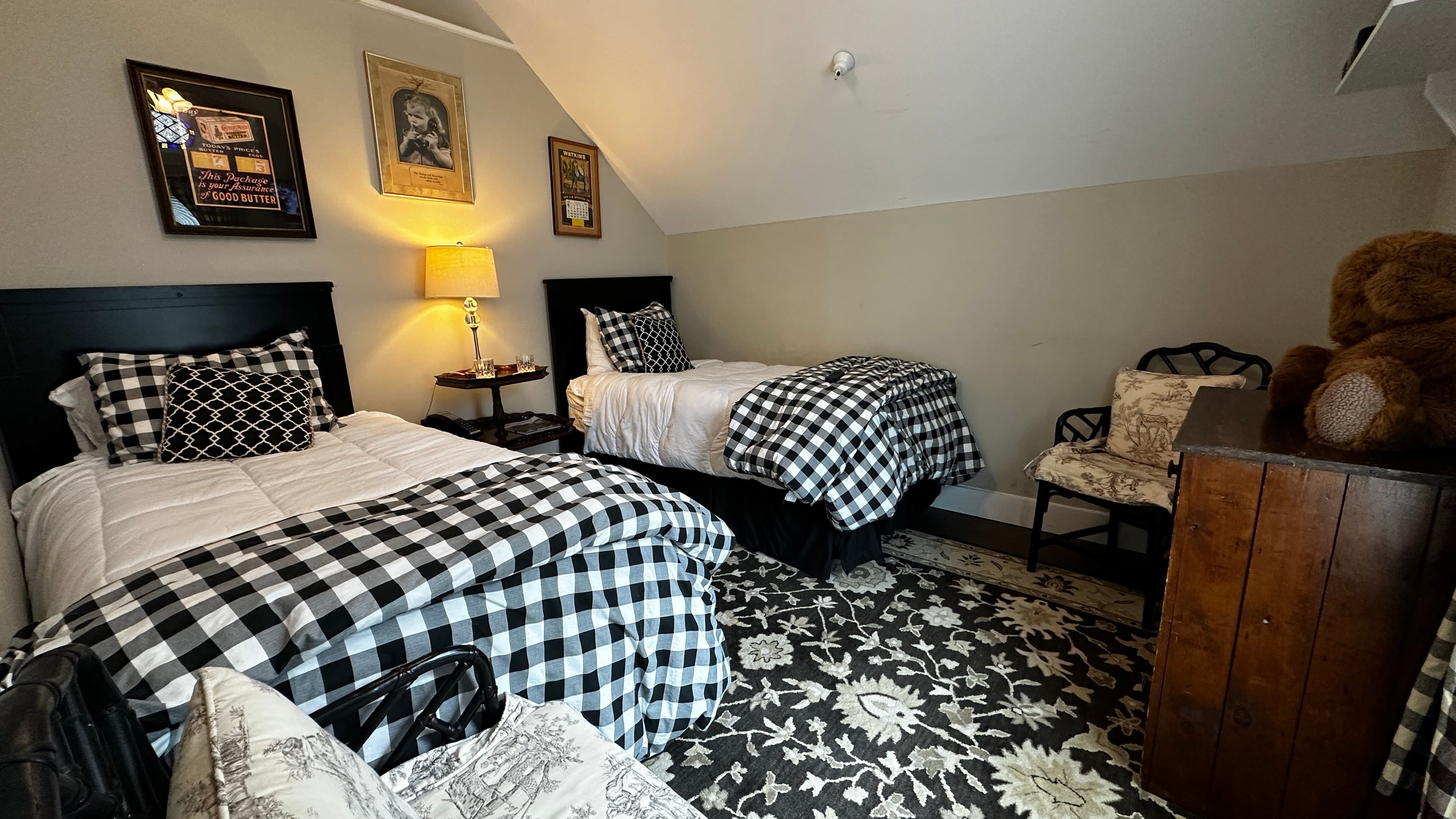 A cozy bedroom with a sloped ceiling, featuring two twin beds with black headboards and buffalo plaid comforters. The room is decorated with a large patterned area rug, vintage-style framed wall art, a warm bedside lamp, and a large teddy bear sitting on a wooden dresser in the foreground.