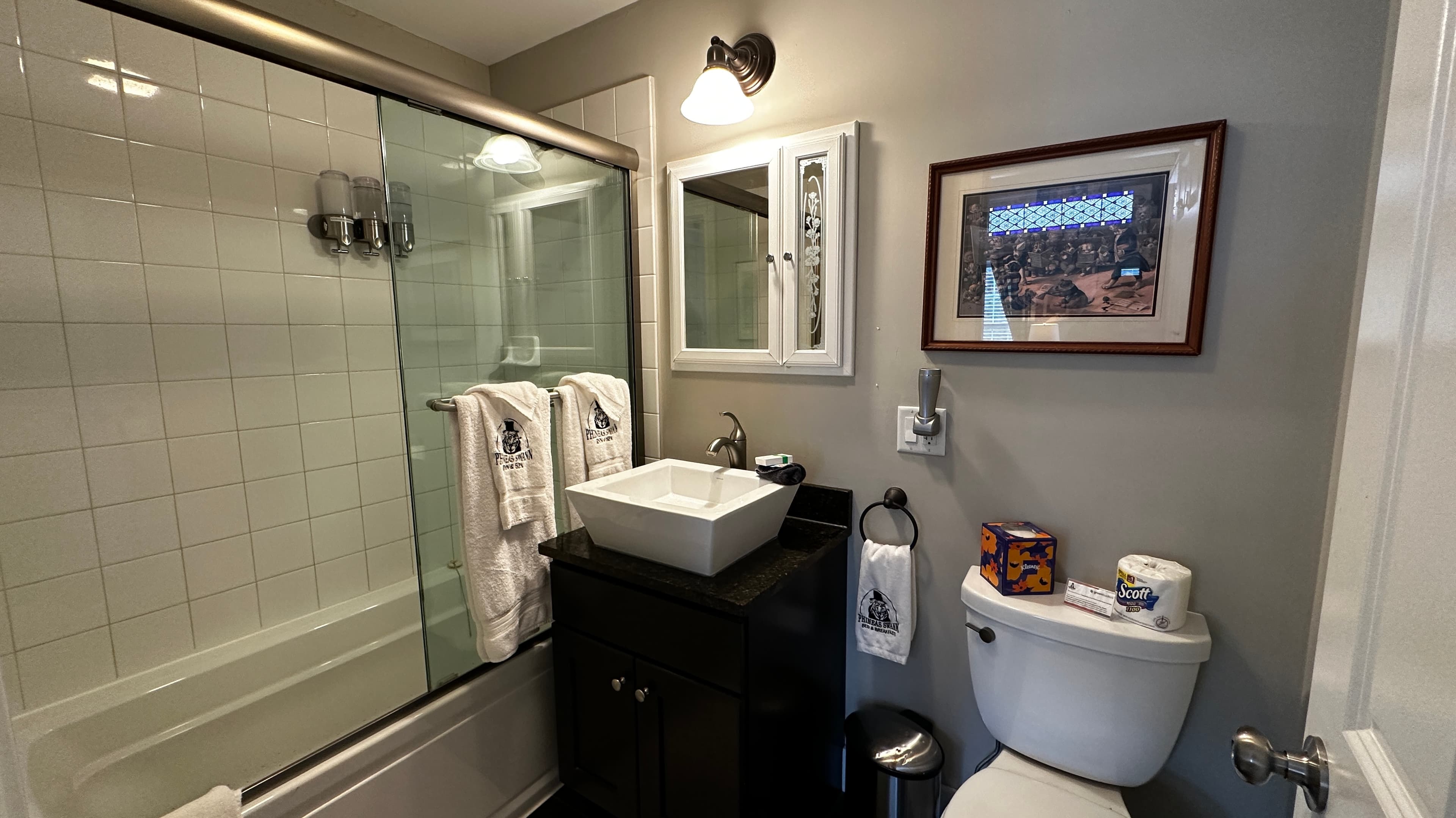 A modern bathroom featuring a white vessel sink on a dark vanity, a white toilet, and a bathtub with sliding glass doors. The room includes a wall-mounted medicine cabinet with a mirror, a framed picture, and neutral grey walls.