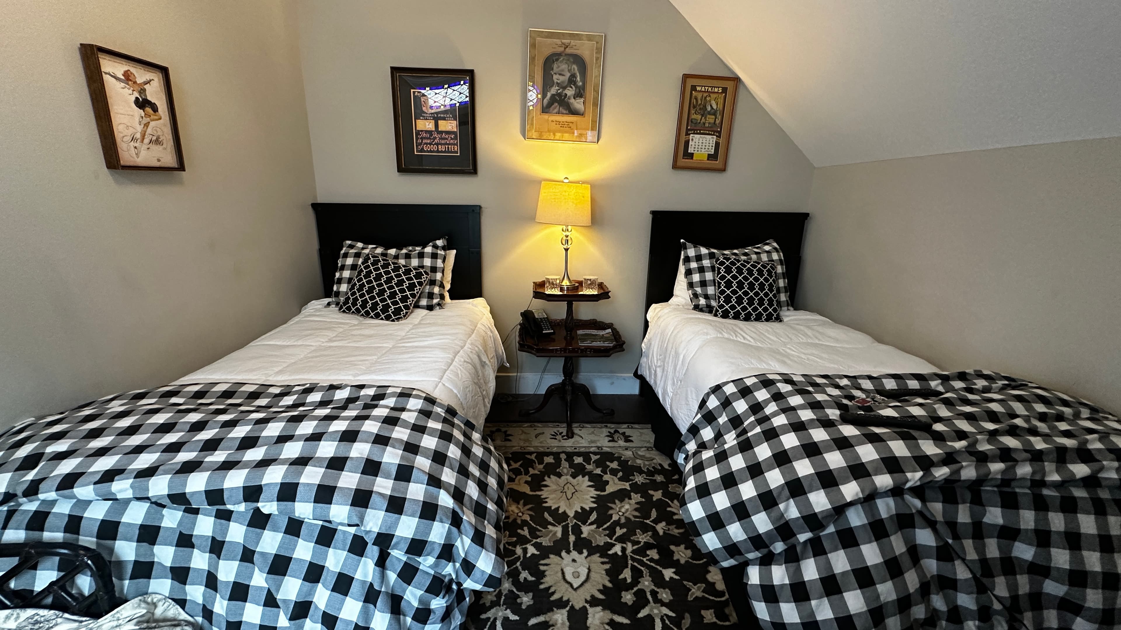 A cozy bedroom featuring two twin beds with black headboards and buffalo plaid comforters. The room is decorated with several framed vintage nature prints on the wall and a central nightstand with a warm lamp casting a soft glow.