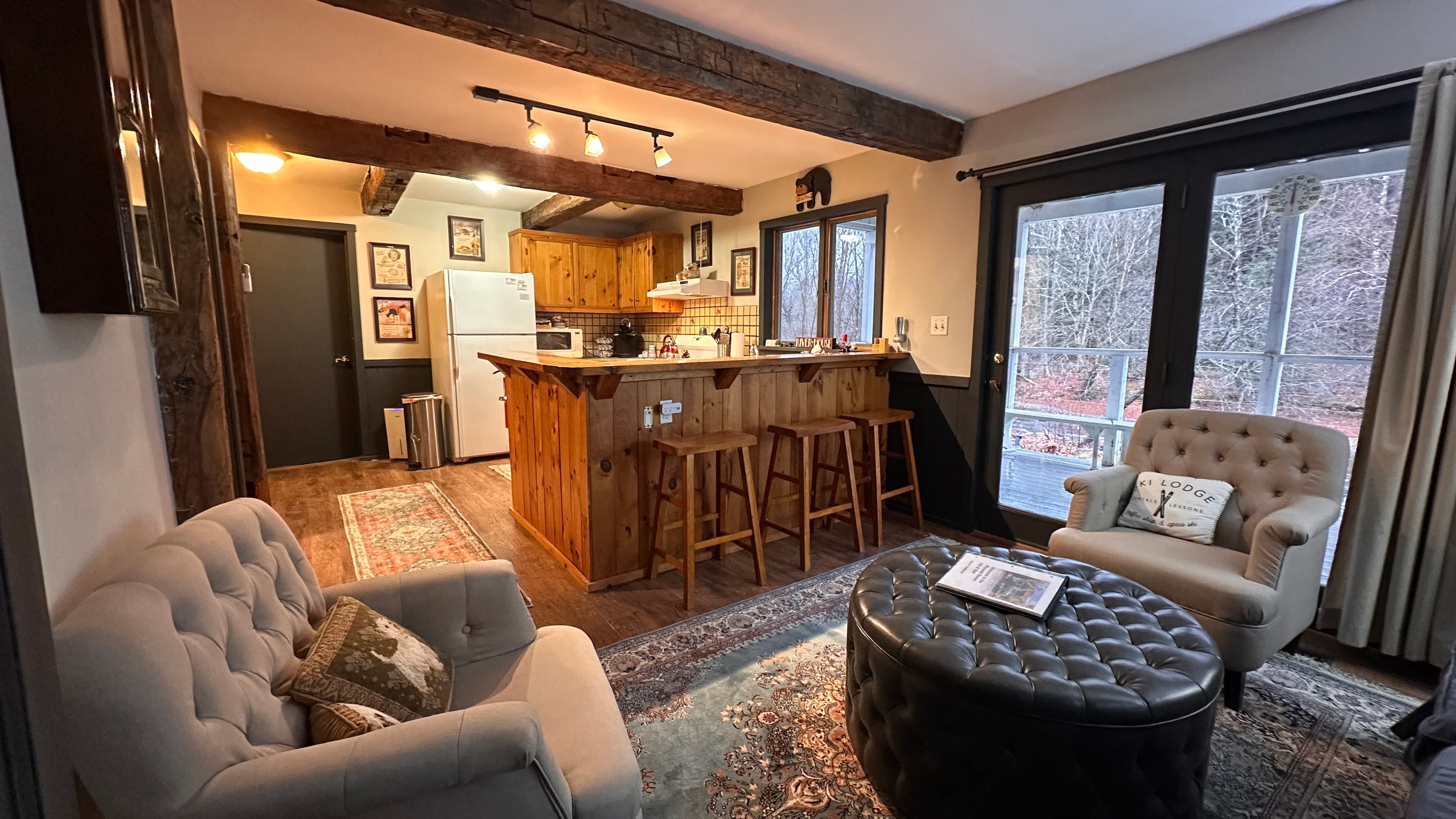 A cozy open-concept living area and kitchen with rustic wooden beams. The space features two grey tufted armchairs, a large black button-tufted ottoman, and a wooden kitchen island with three barstools. In the background, a small kitchen with white appliances is visible, along with a glass door leading to a wooden deck..