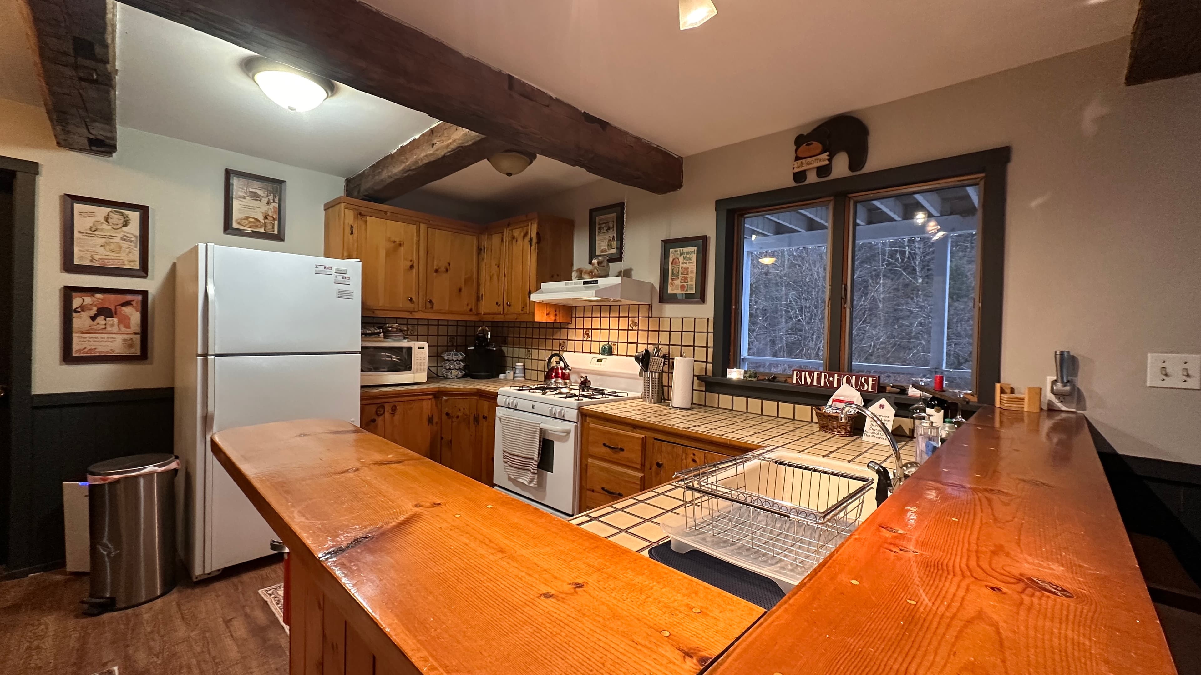A rustic kitchen featuring warm wood cabinetry, white appliances, and a large wooden breakfast bar. The space is accented by dark exposed ceiling beams, track lighting, and framed artwork on the walls, with a window overlooking a wooded outdoor area.