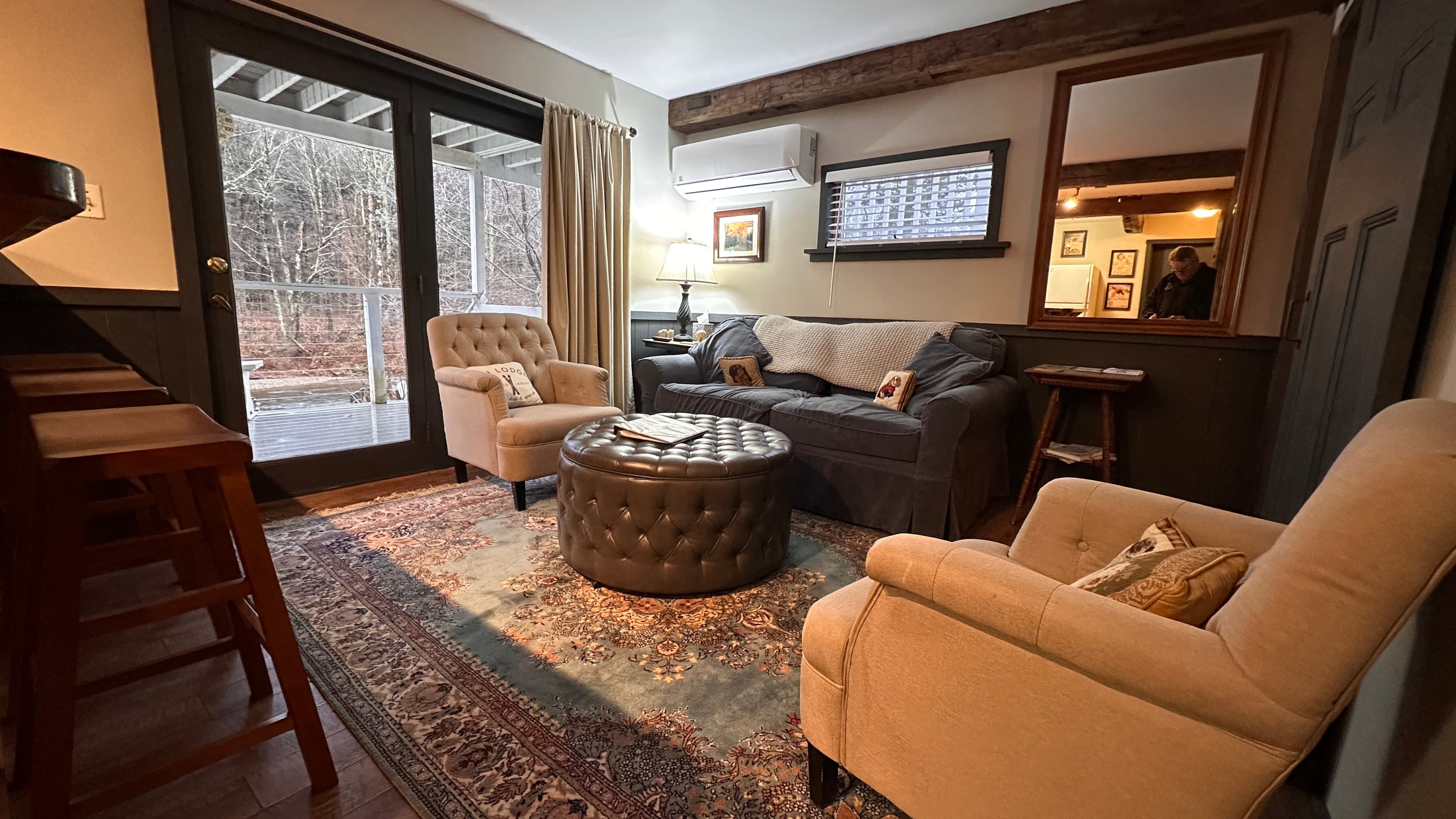 A warm and inviting living area with a gray sofa, two cozy beige armchairs, and a tufted ottoman. Glass doors lead to a wooden deck.