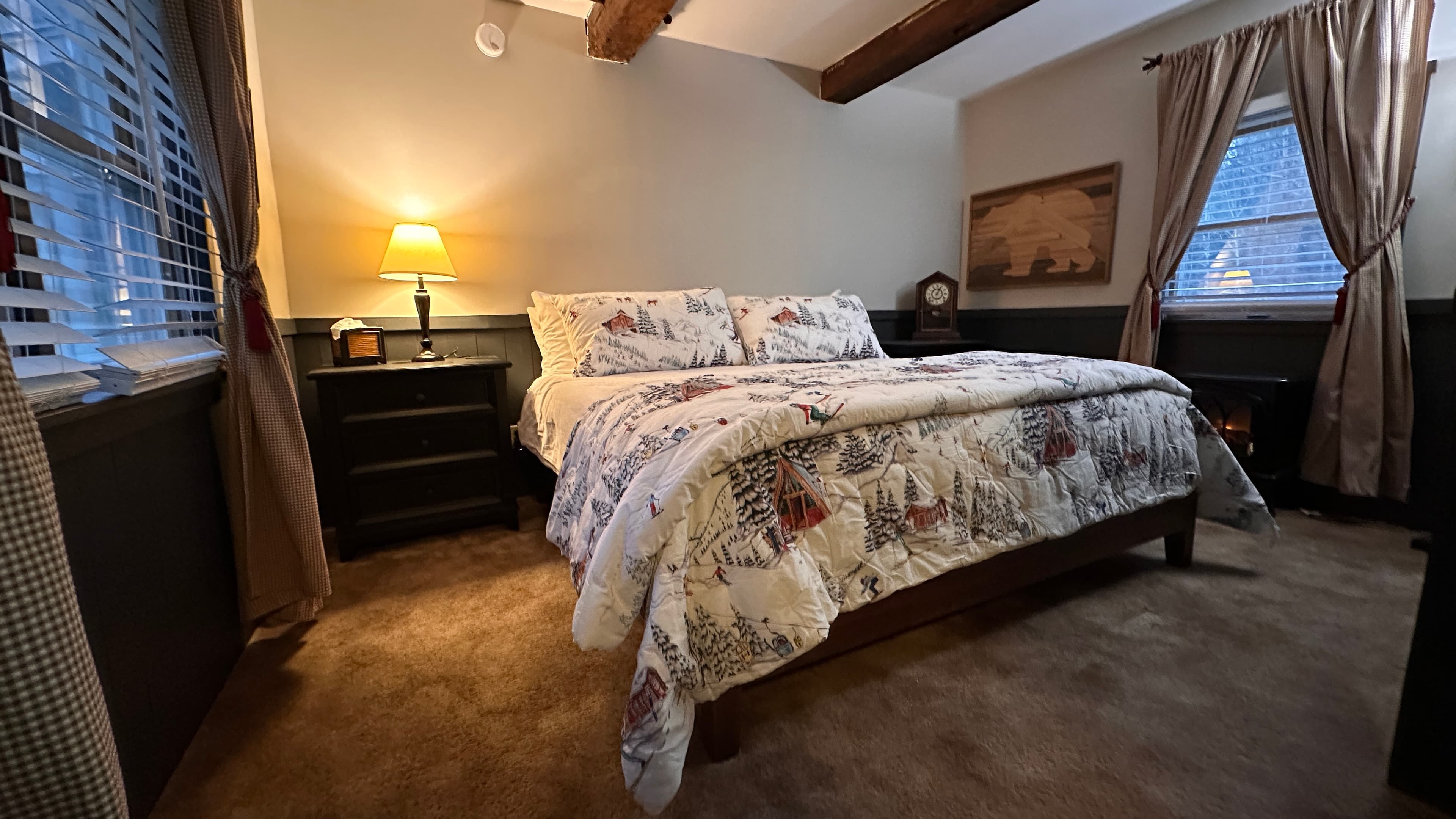 A cozy, rustic bedroom featuring a bed with a nature-patterned comforter, exposed wooden ceiling beams, and warm lamp lighting on a dark wood nightstand.