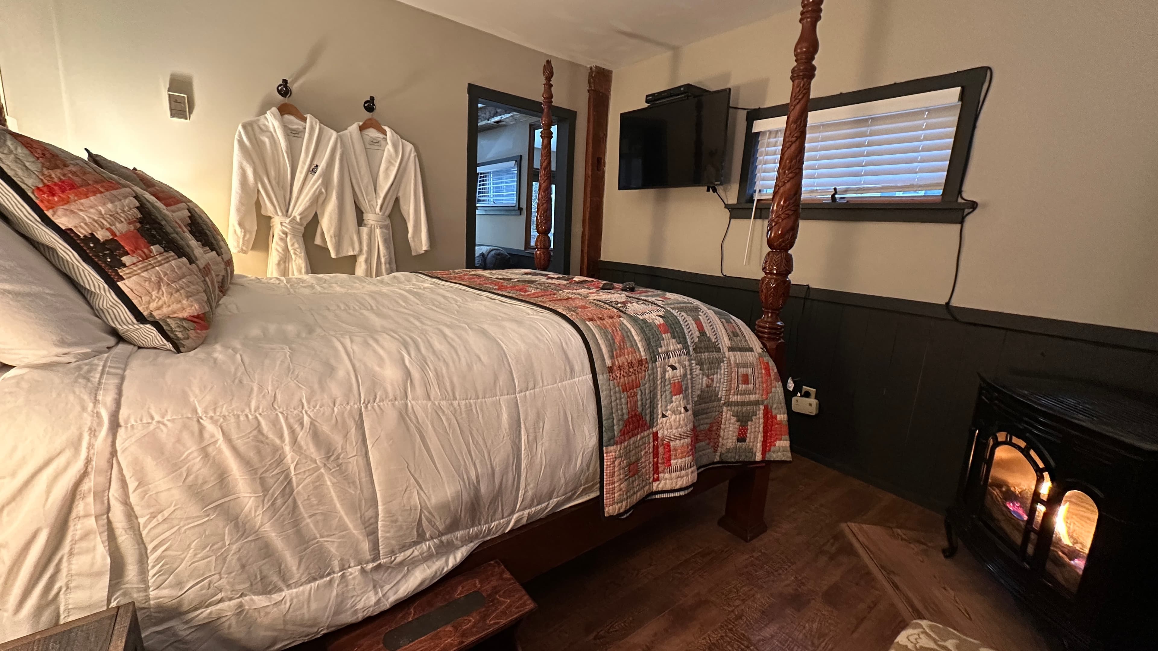 A bedroom with a four-poster bed featuring a white comforter and patterned accent pillows, two white robes hanging on the wall, and a glowing fireplace in the corner.
