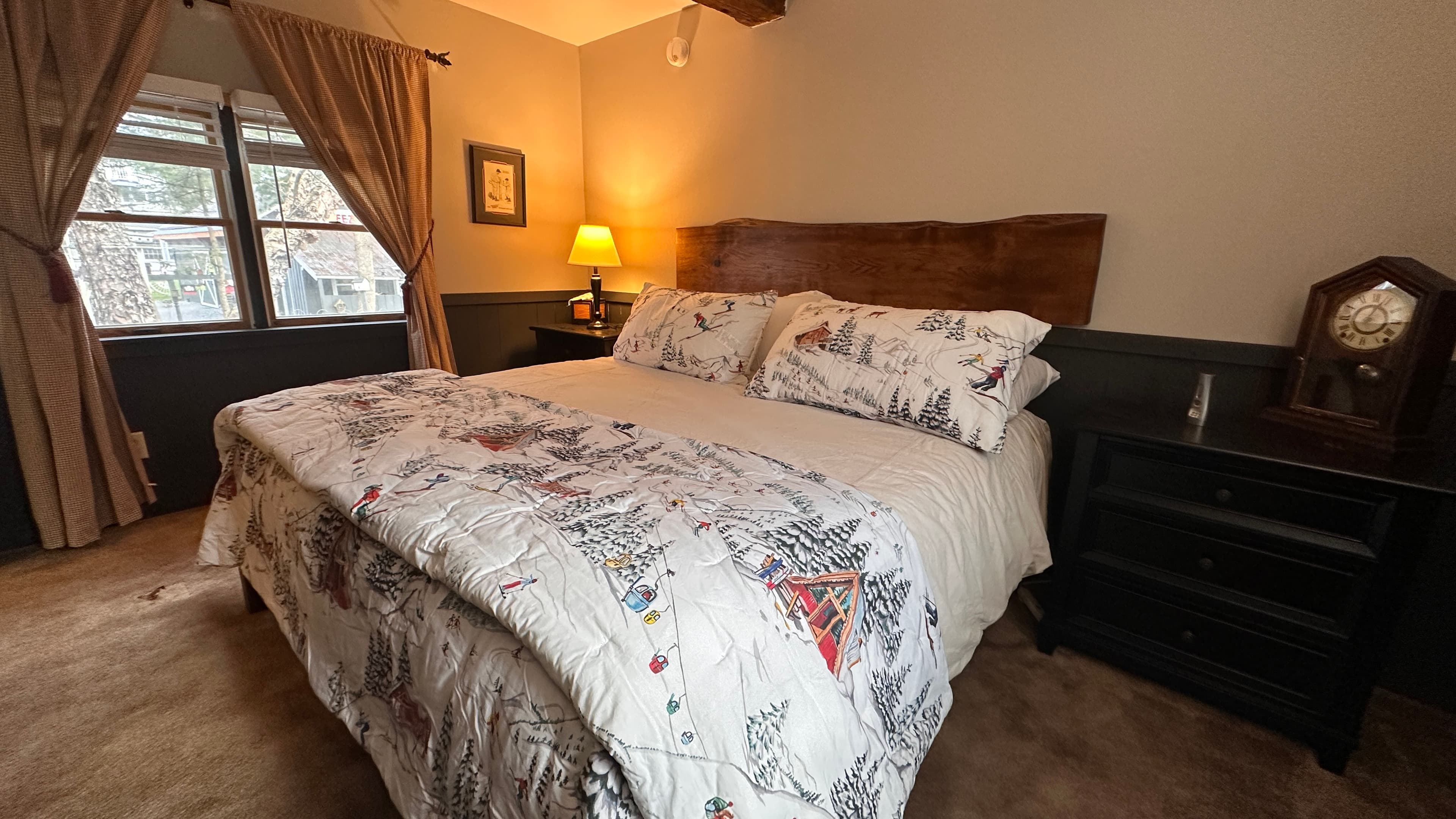 A cozy bedroom featuring a wooden king-sized bed with a live-edge headboard and white bedding with a winter-themed pattern. The room includes a black bedside table with a vintage-style clock, a warm lamp, and a large window with tan curtains overlooking a wooded area.