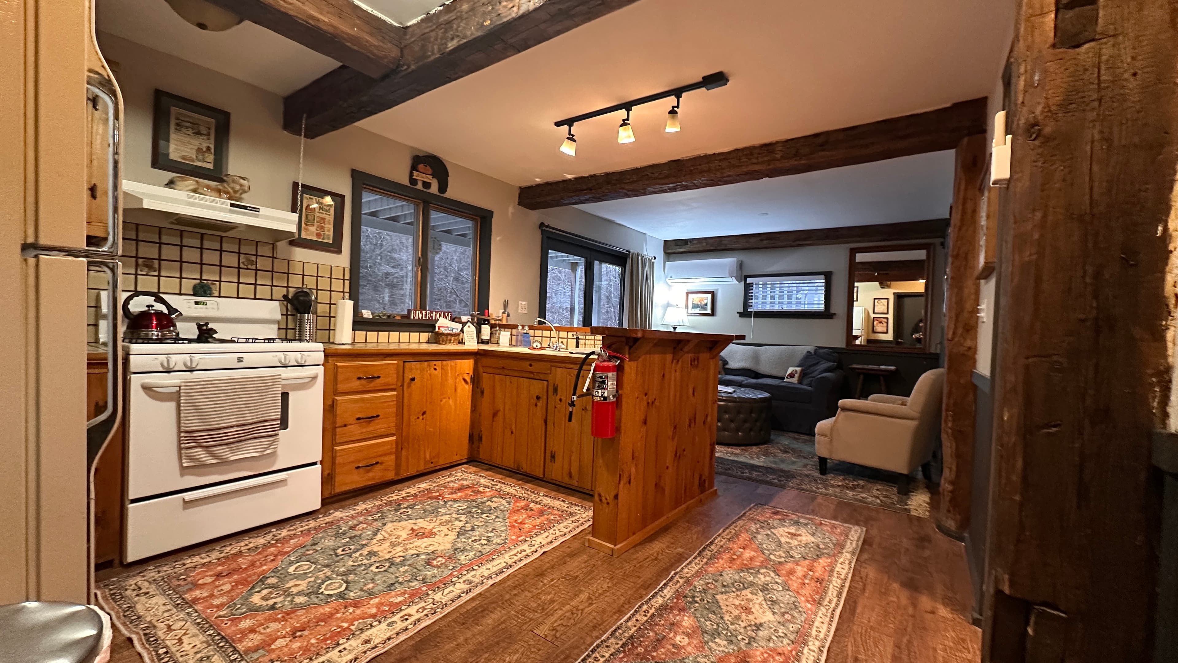 A cozy, rustic kitchen and living area featuring dark exposed wooden ceiling beams and warm track lighting. The kitchen includes white appliances, natural wood cabinetry, and a peninsula with barstools, while the adjacent living space offers grey tufted armchairs and a large black button-tufted ottoman on a patterned area rug.