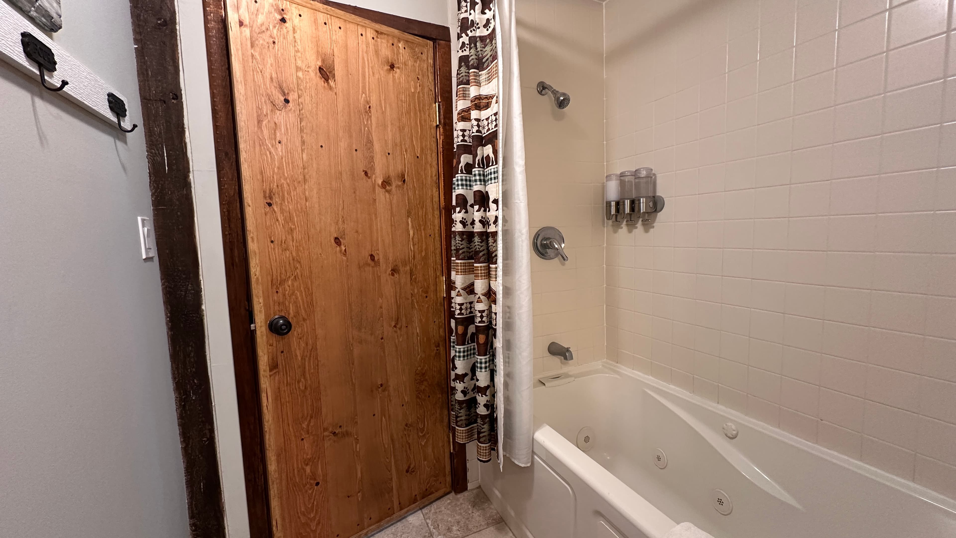 A cozy bathroom featuring a large white jetted bathtub with a showerhead and a patterned shower curtain. The room includes a rustic wood-plank door, white tiled walls, and wall-mounted dispensers for toiletries.