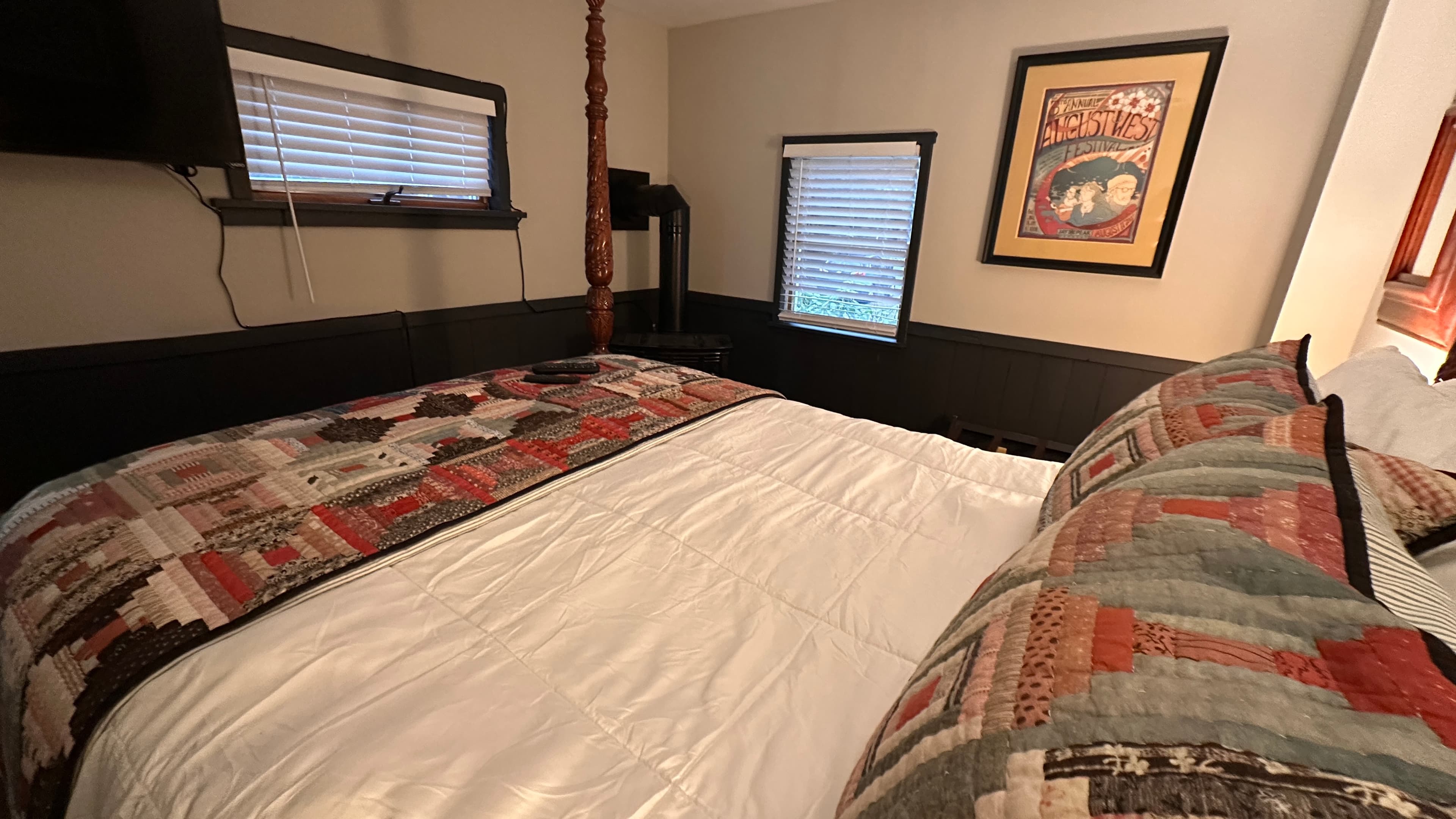 A cozy bedroom featuring a dark wood four-poster bed with white linens and a Southwestern-patterned quilt runner. The room is decorated with dark grey wainscoting, framed wall art, and small windows with white blinds, creating a warm and rustic atmosphere.