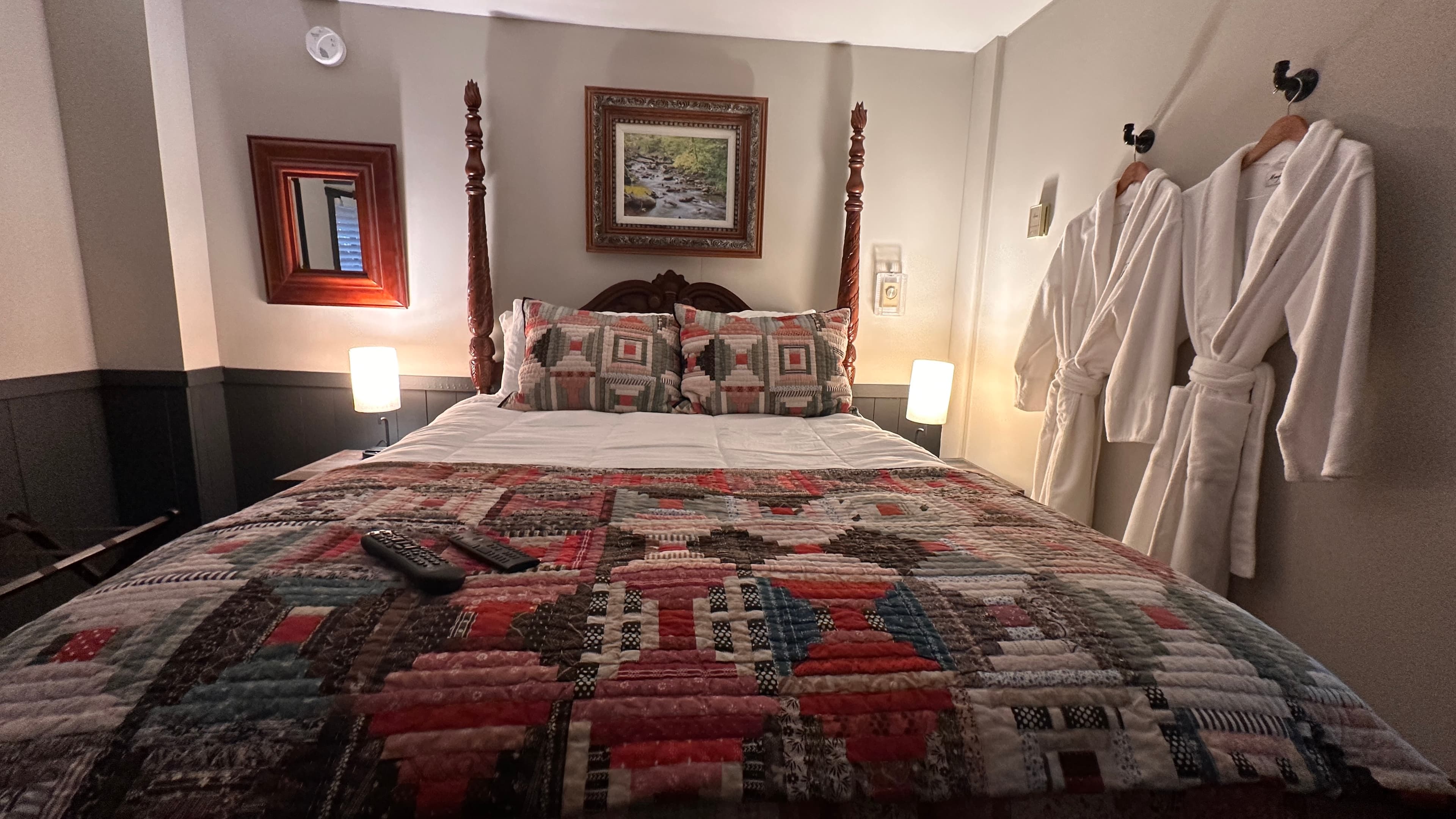 A cozy bedroom featuring a dark wood four-poster bed with white linens and a Southwestern-patterned quilt runner. The room is decorated with dark grey wainscoting, framed wall art, and small windows with white blinds, creating a warm and rustic atmosphere.