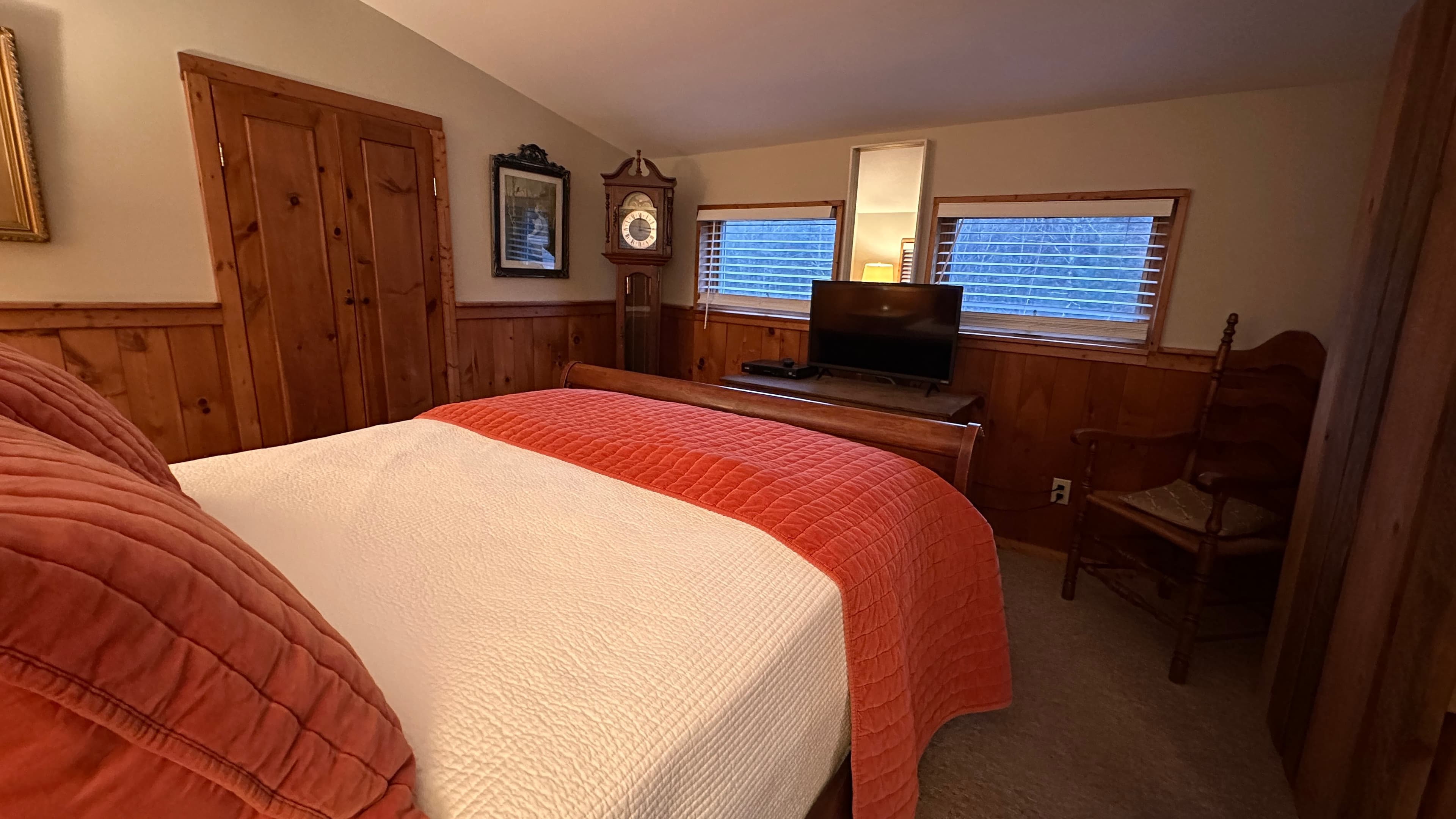 A cozy bedroom featuring a bed with white linens and a salmon-colored textured blanket and pillows. The room has warm partially wood-paneled walls, two small windows, a vintage-style standing clock, and a classic wooden chair in the corner.