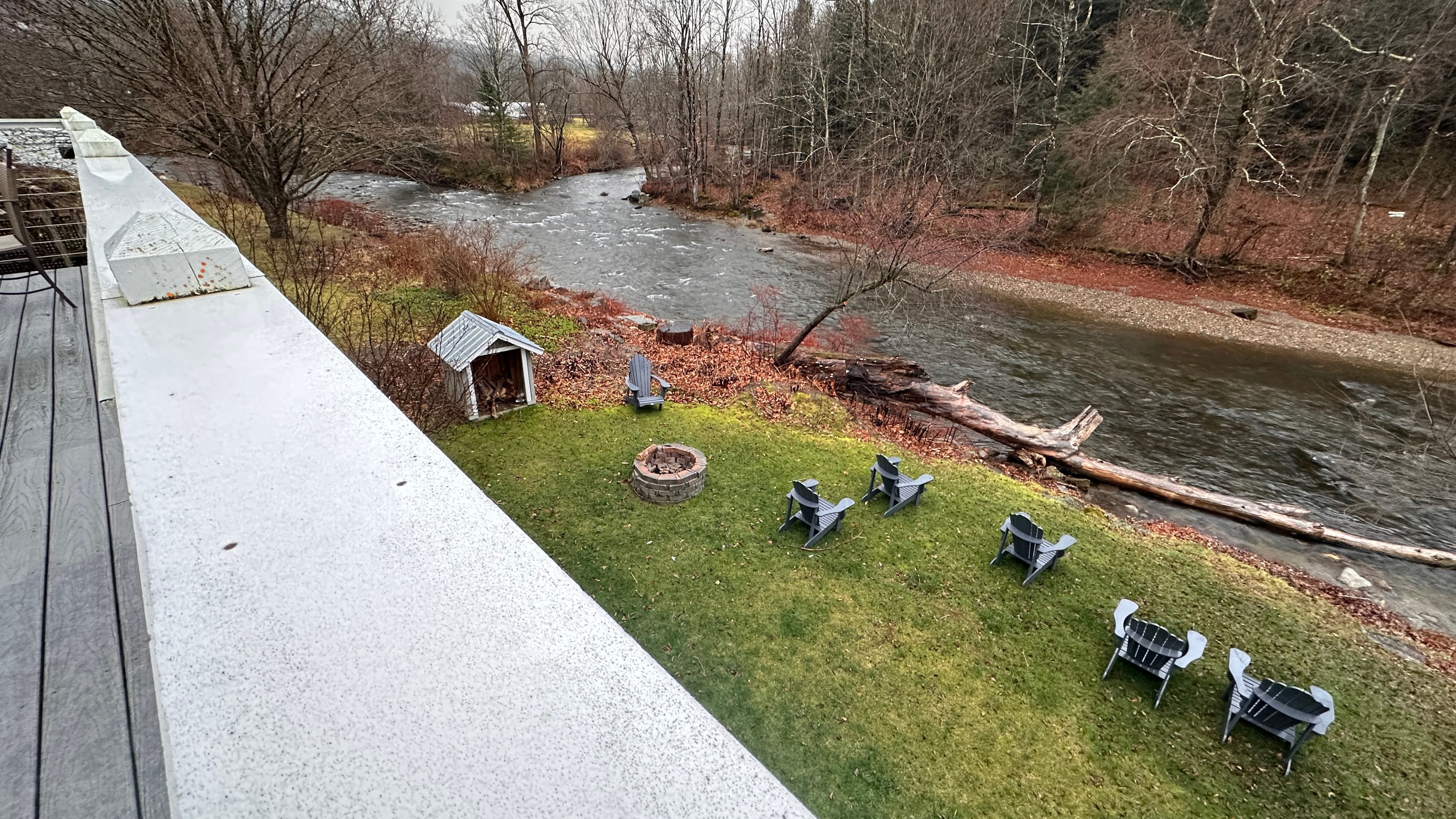 A view from the deck of a grassy backyard bordering a flowing river. The yard features several black Adirondack chairs arranged facing the water and a stone fire pit.