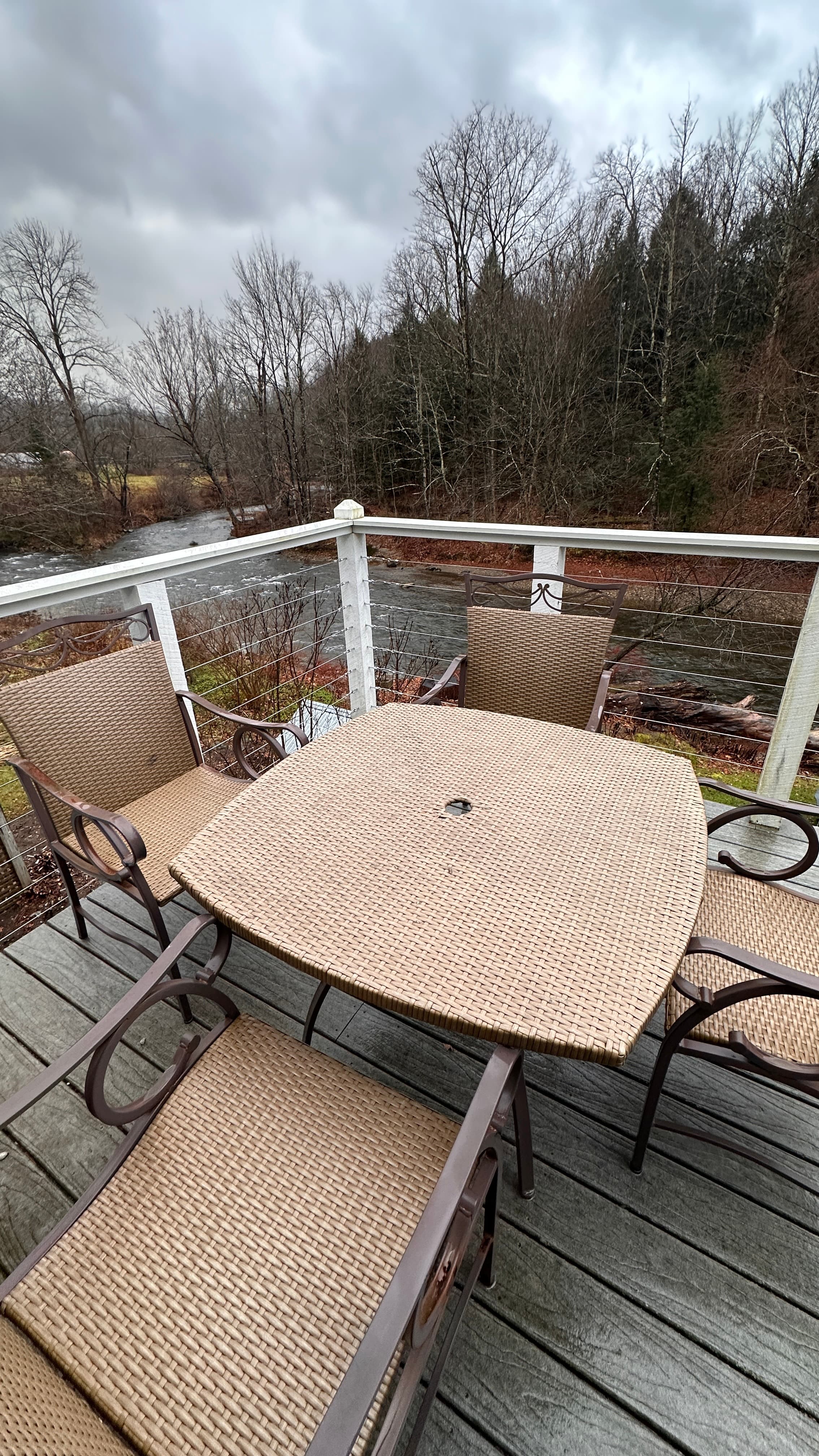 A high-angle view from a wooden deck looking down onto a patio set with a square tan table and four matching mesh chairs. In the background, a river flows past a grassy bank and a dense forest under a cloudy sky.