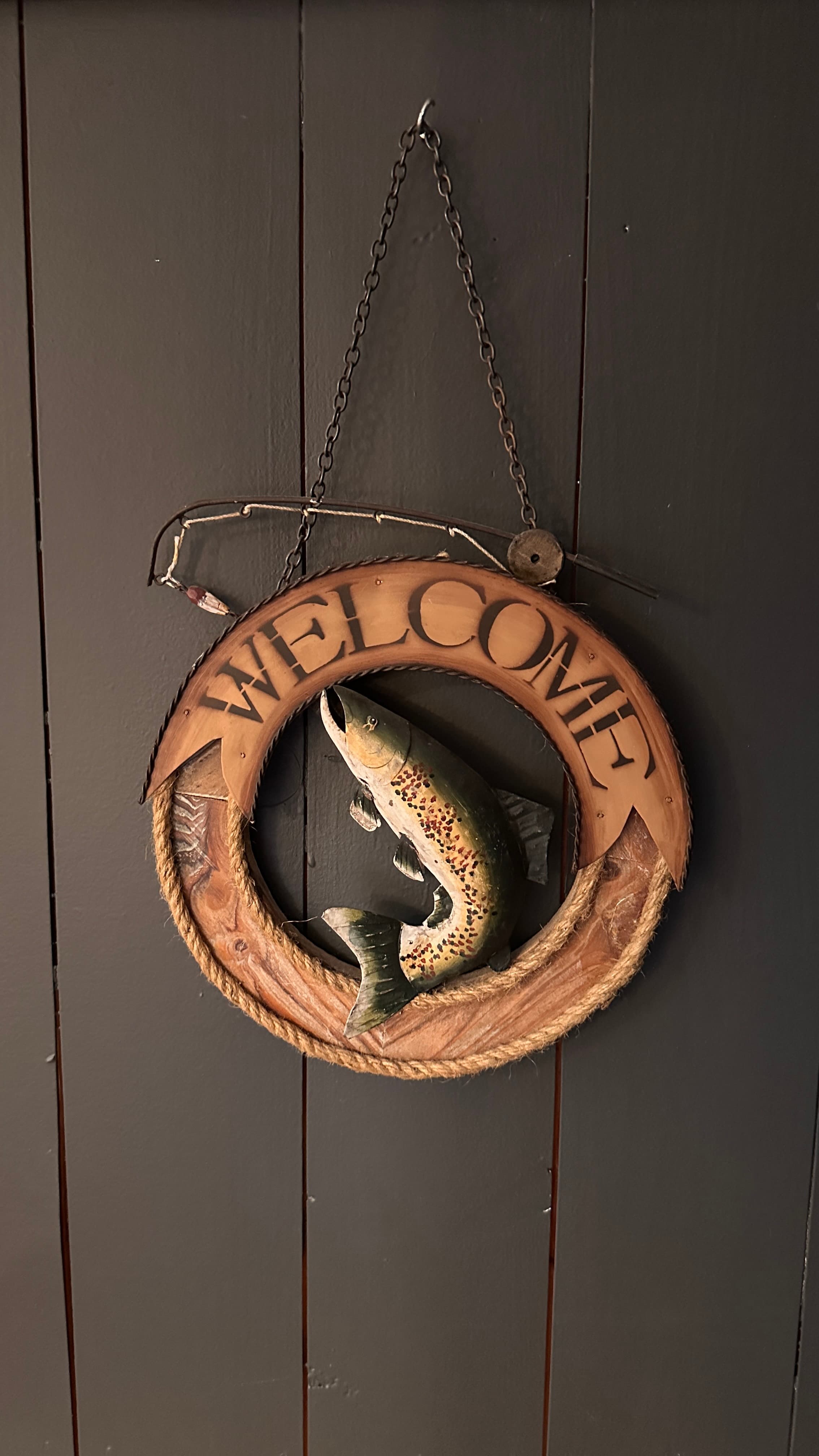A circular wooden "Welcome" sign hanging from a chain on a dark, vertical-paneled wall. The sign features a carved fish jumping in the center and a small decorative fishing rod along the top edge.