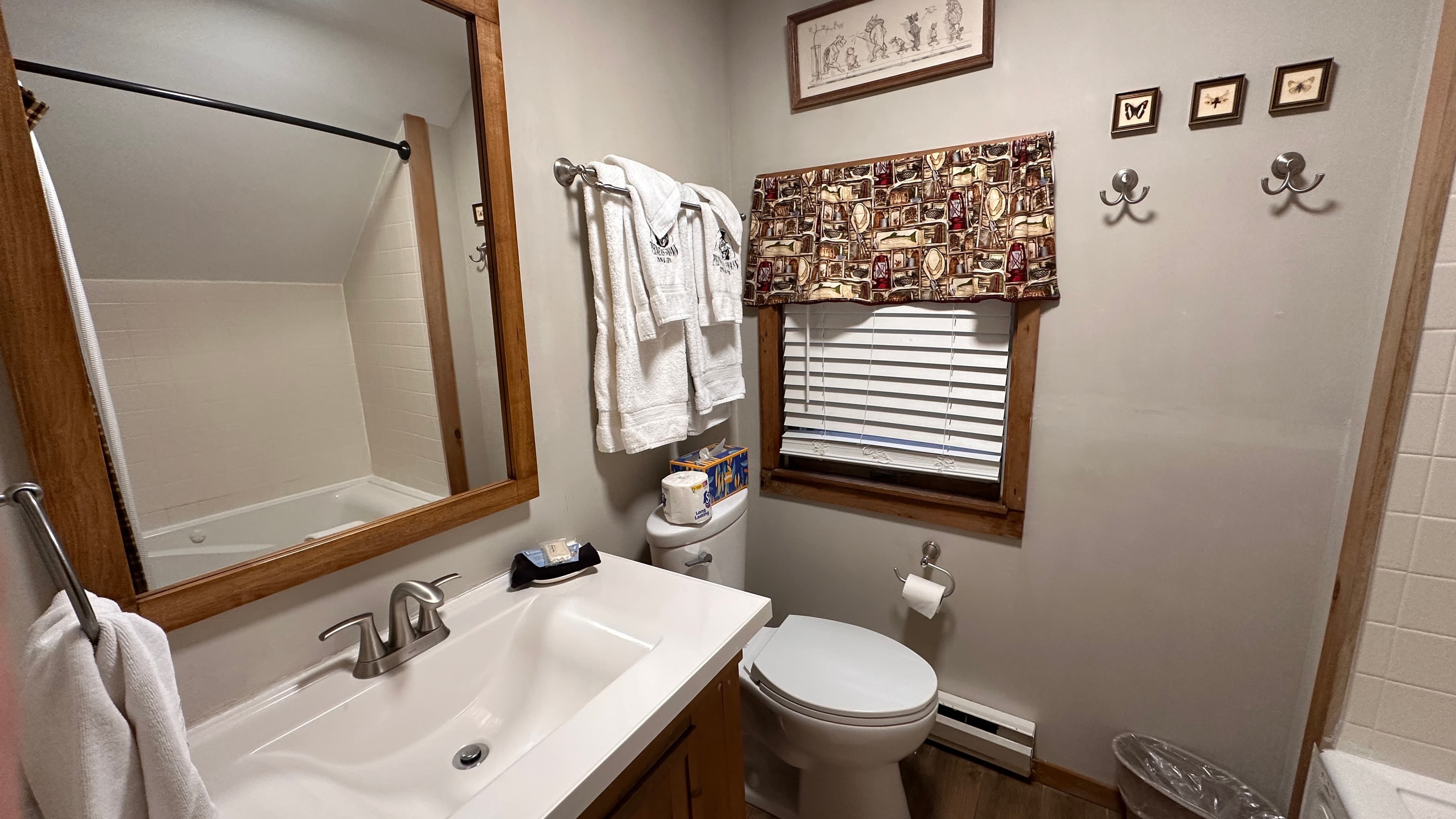 A small, rustic-style bathroom featuring a white vanity with a chrome faucet, a large wood-framed mirror reflecting a bathtub, and a toilet. The wall is decorated with a patterned valance over a window, framed art, and decorative wall hooks.