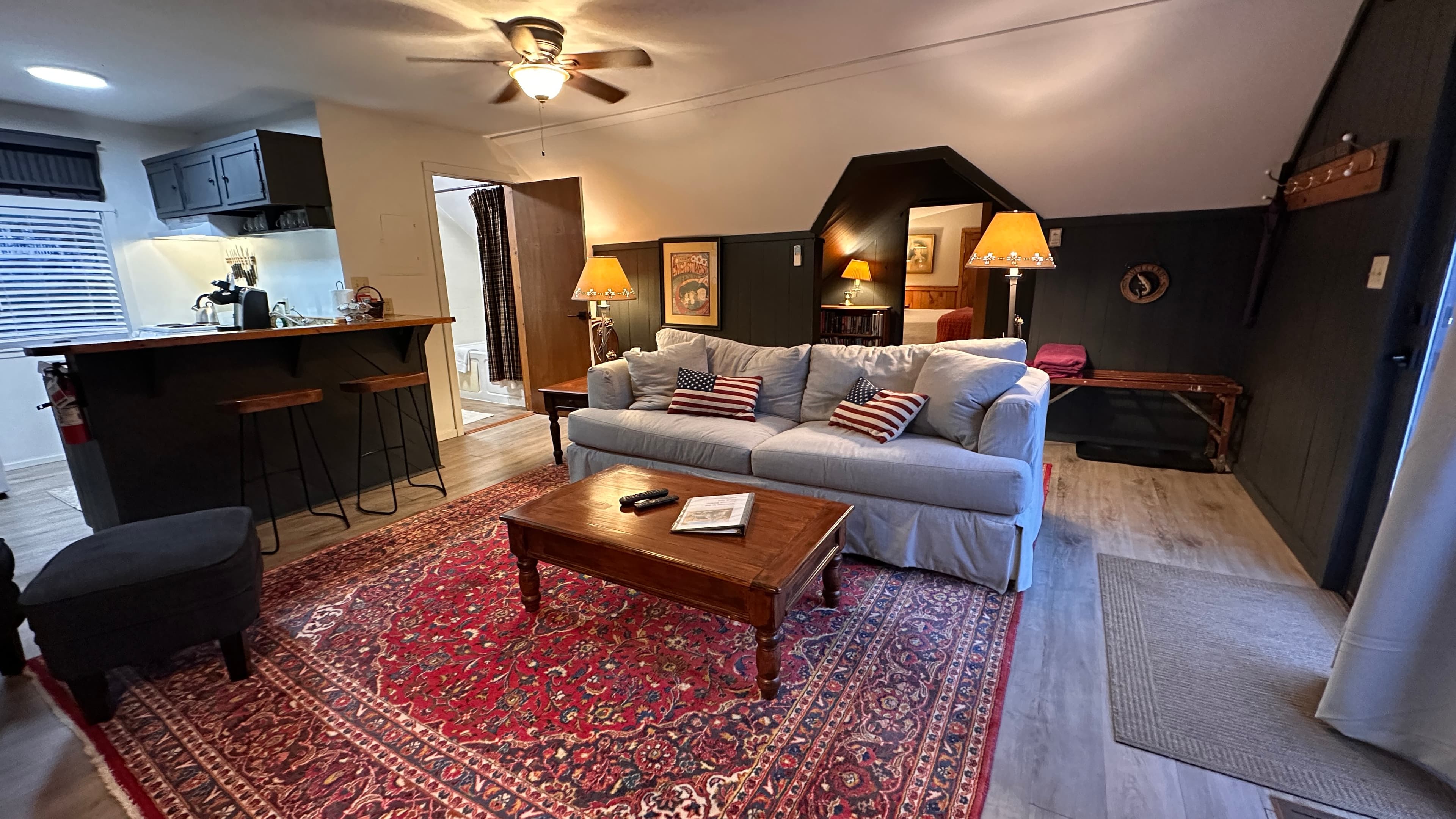 A living room featuring a light gray sofa and a wooden coffee table centered on a large red patterned rug. In the background, a dark wood breakfast bar with stools separates the living area from a small kitchen, and a doorway leads to another room.
