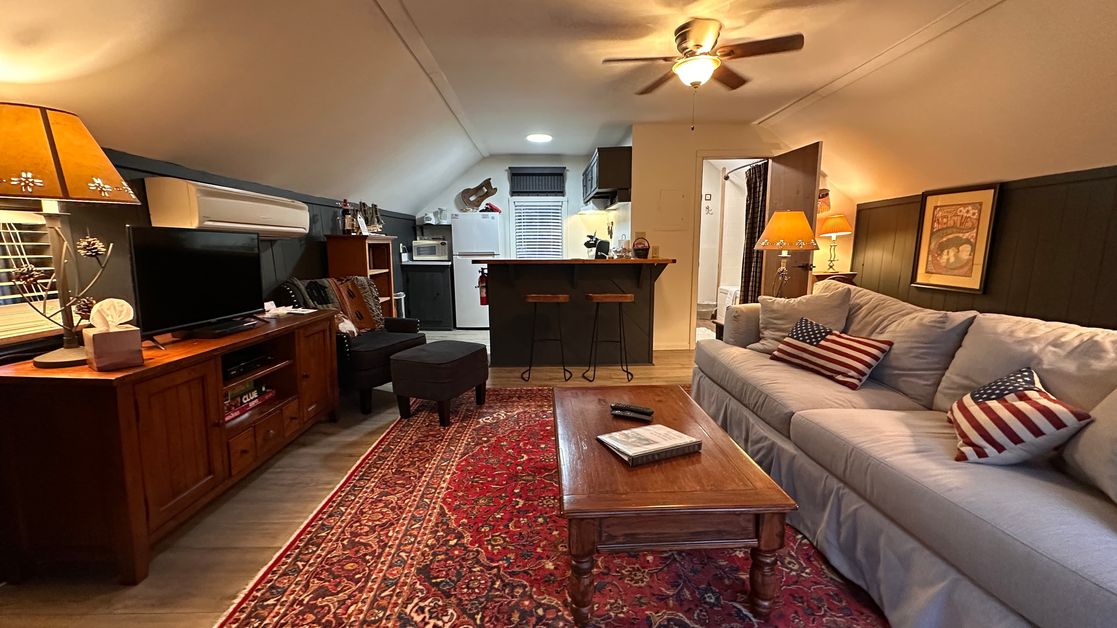 A cozy living area featuring a light gray sofa with decorative pillows and a wooden coffee table centered on a large red patterned rug. In the background, a dark wood breakfast bar separates the living space from a small kitchen, with a hallway leading to other rooms under a slanted white ceiling with a ceiling fan.