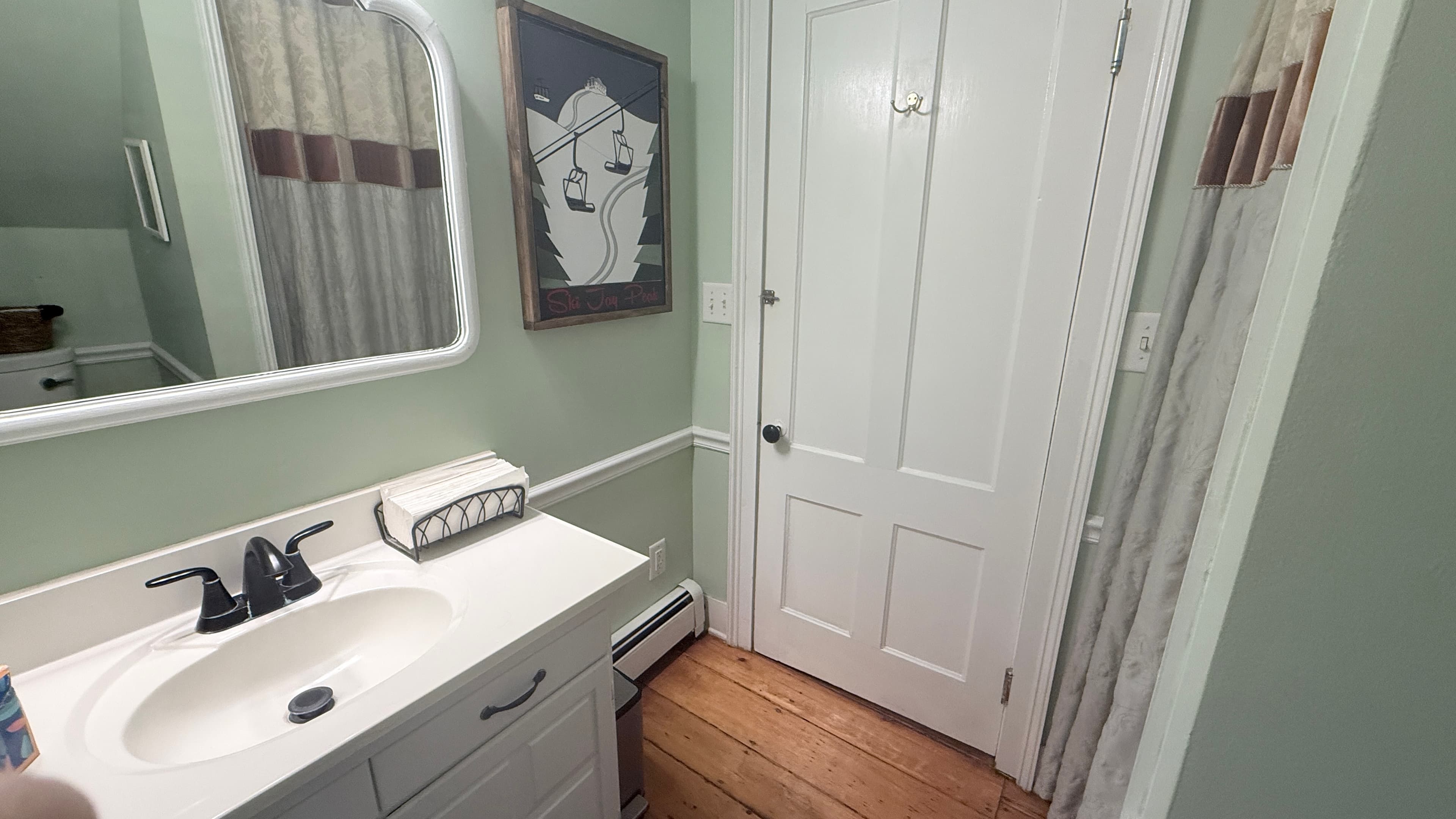 A view of a bathroom with pale green walls and white wainscoting, featuring a white vanity with a dark faucet and a rounded mirror. To the right, a white door stands closed next to a shower area with a tan and white patterned curtain. A framed piece of art hangs on the wall between the mirror and the door.