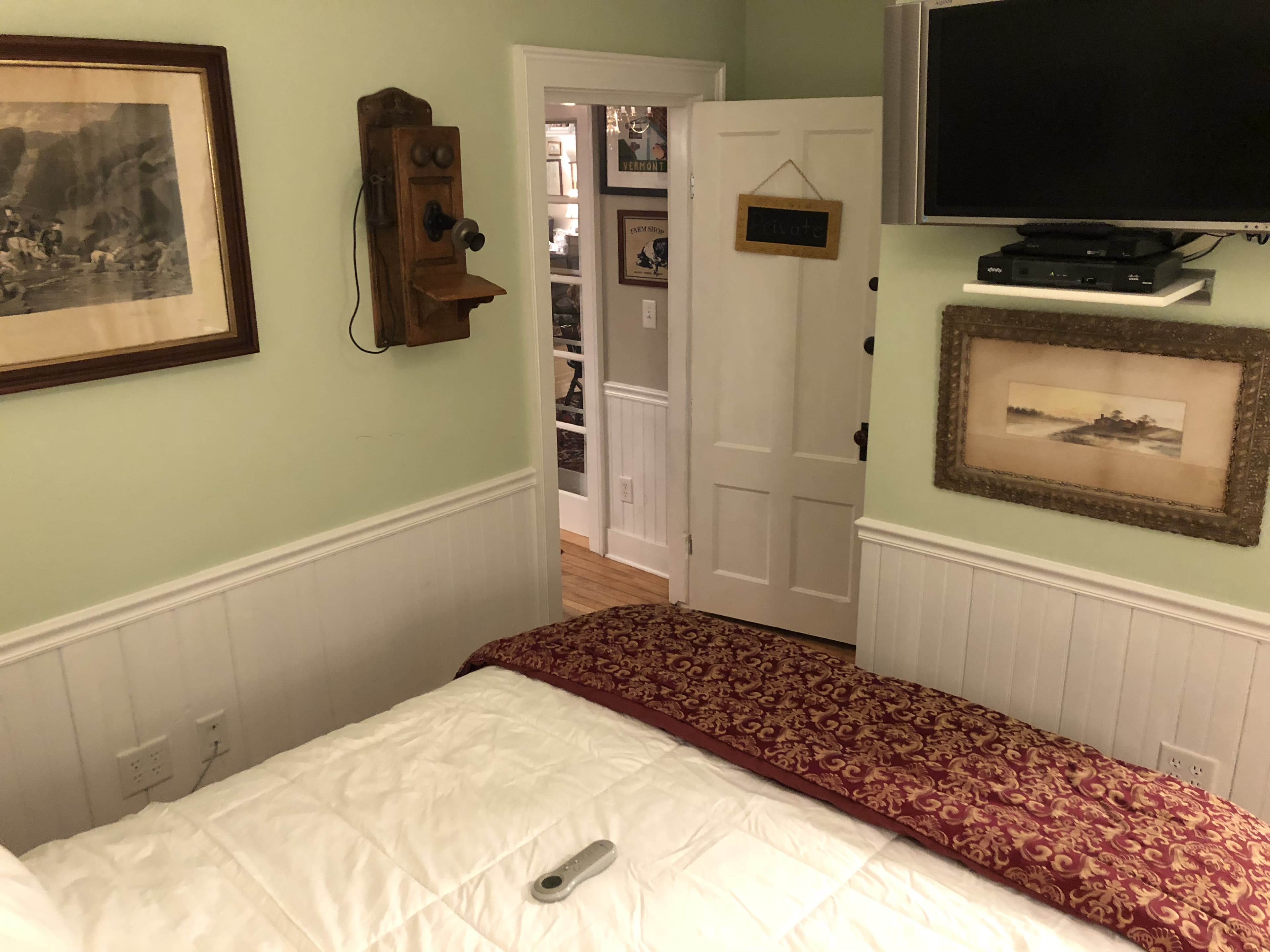 A cozy bedroom with pale green walls and white wainscoting. A bed with white linens and a dark patterned throw occupies the foreground, while the wall features a mounted television, framed artwork, and a vintage-style wooden wall phone. An open white door leads into another room with a bookshelf.