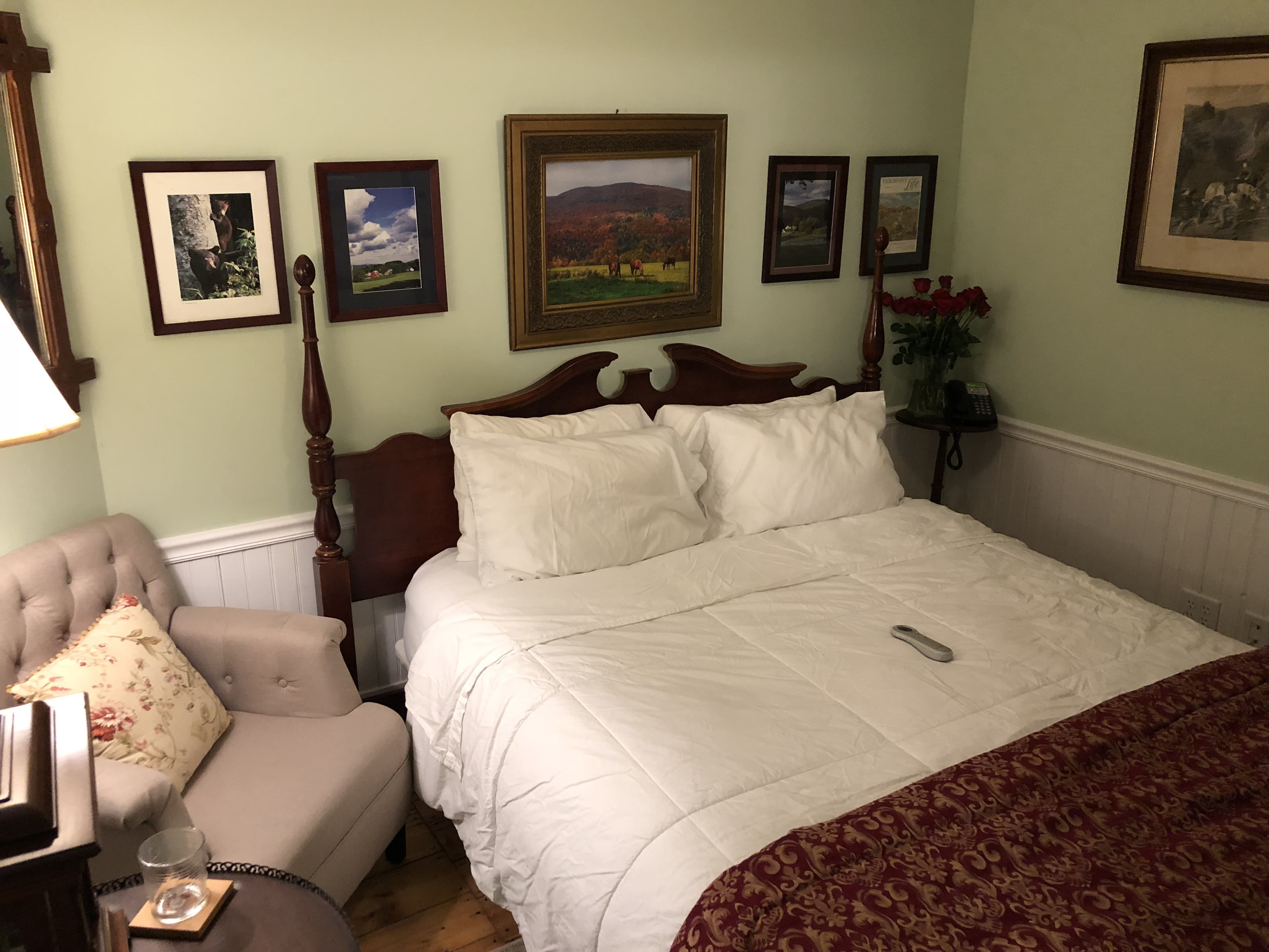 A high-angle view of a cozy bedroom with light green walls and white wainscoting. The bed is neatly made with white linens and a dark, patterned red throw blanket. The room is decorated with a gallery wall of framed landscape art above the dark wood headboard, a tufted armchair in the corner, and a television mounted on the wall above a framed picture.