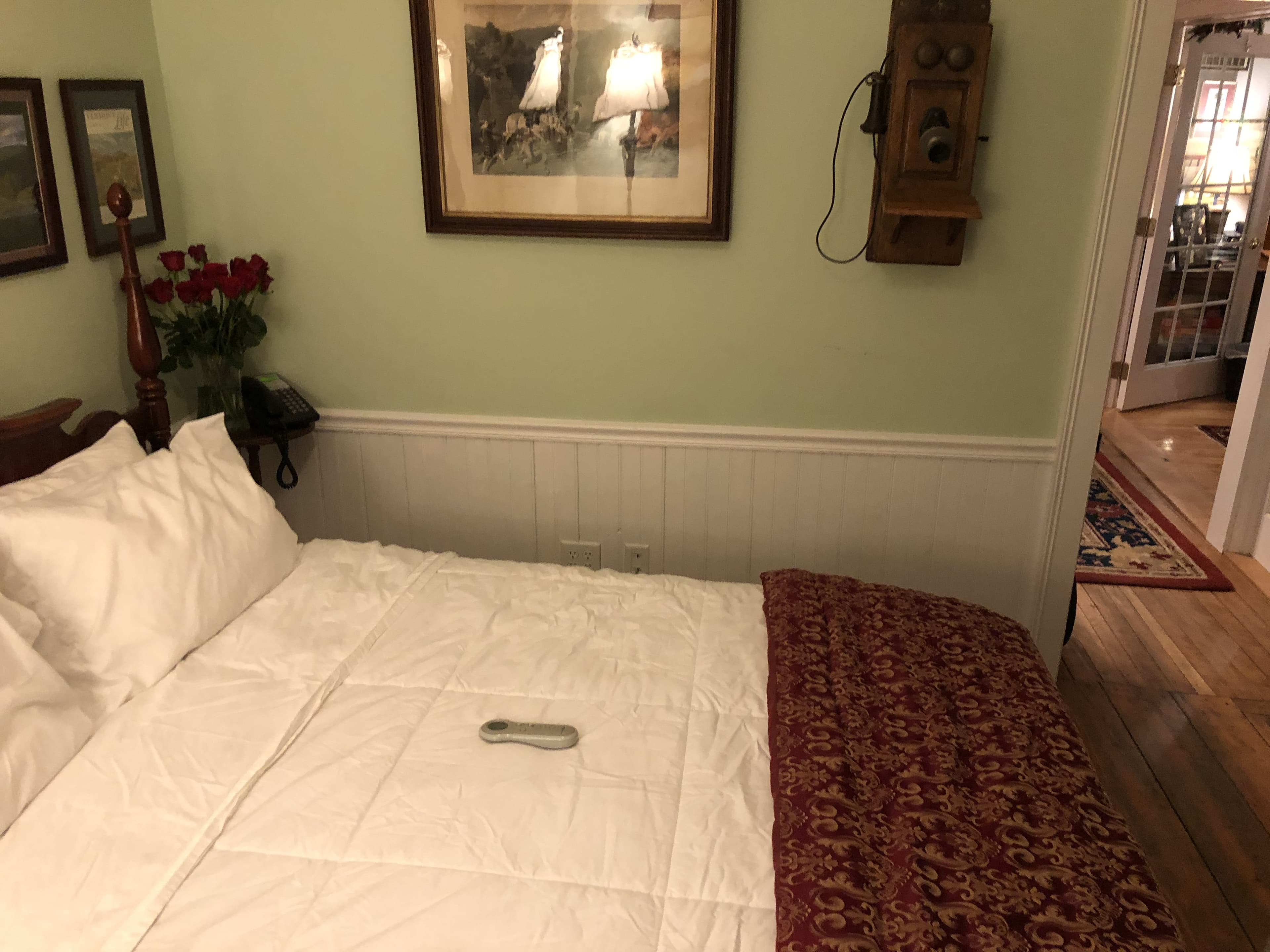 A cozy bedroom featuring a neatly made bed with white linens and a dark red patterned throw. The pale green walls are decorated with a collection of framed landscape art above a dark wood headboard, a vintage-style wooden wall phone, and a television mounted above a framed picture. A tufted armchair sits in the corner, and an open white door reveals a glimpse of a hallway.