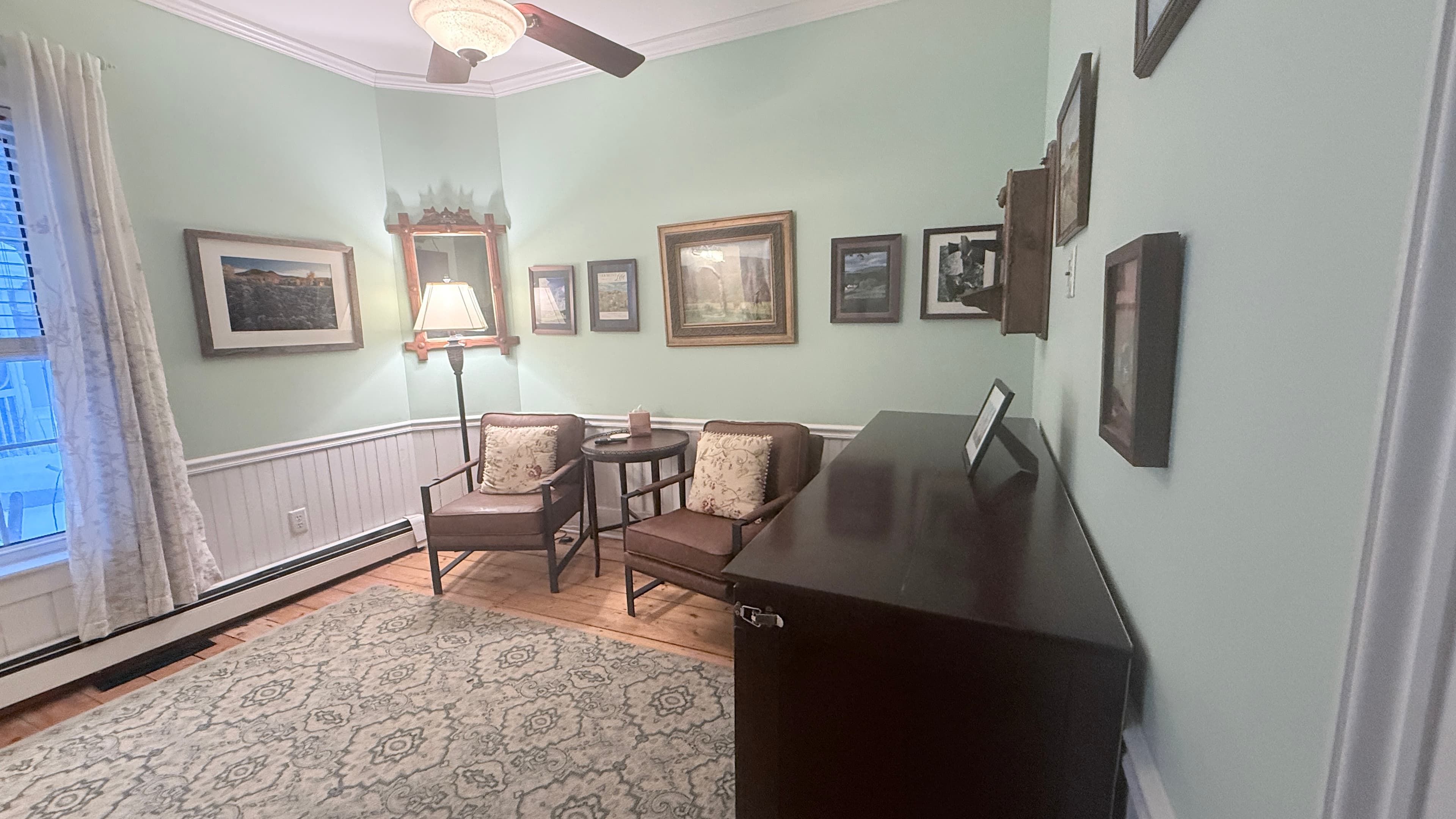A cozy bedroom featuring light green walls, white wainscoting, and a dark wood dresser in the foreground. Two brown armchairs sit against the far wall near a tall lamp and a large window with white curtains. A patterned area rug covers the hardwood floor, and the walls are decorated with a collection of framed landscape art.