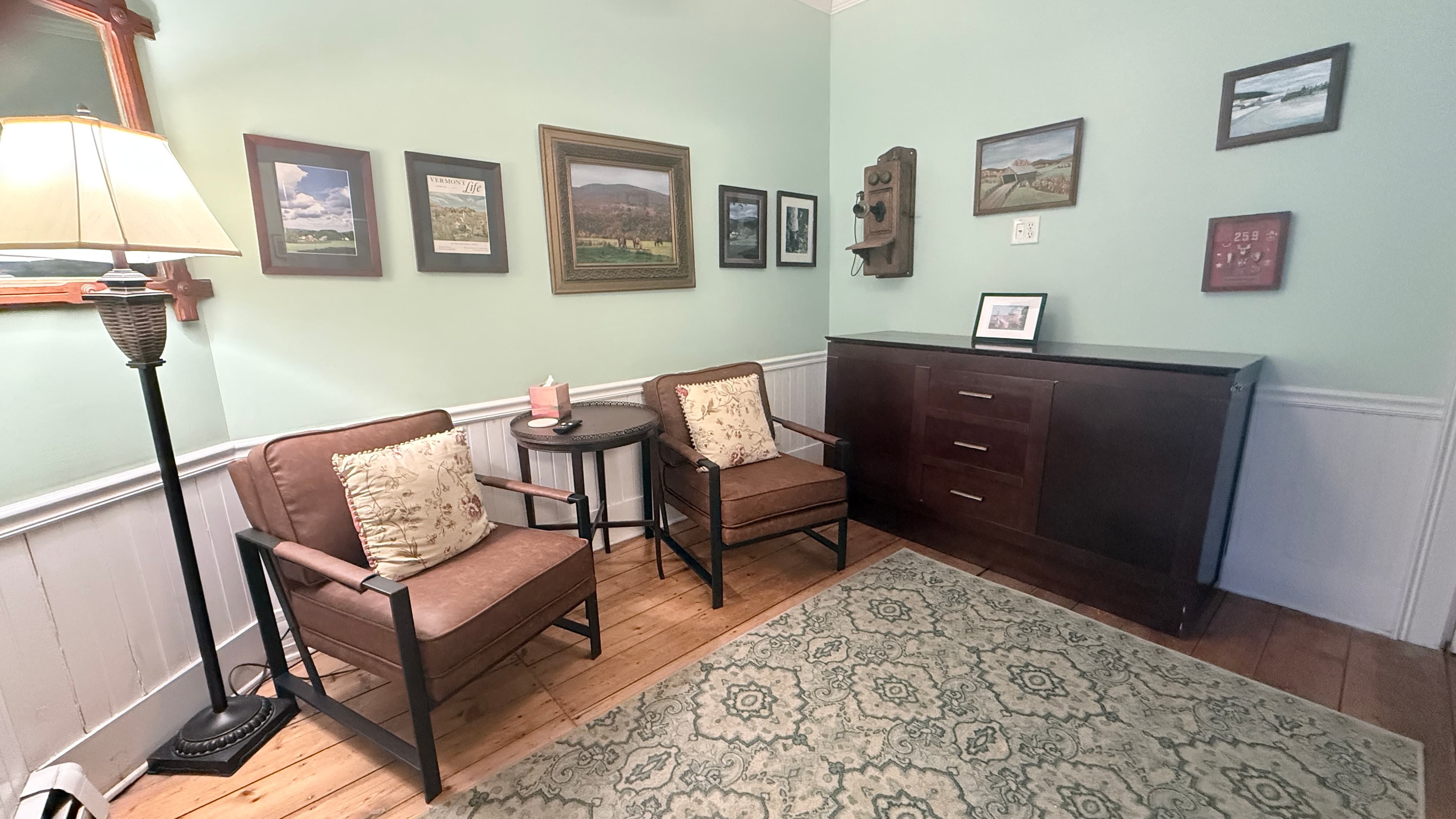 A cozy bedroom featuring light green walls, white wainscoting, and a dark wood dresser in the foreground. Two brown armchairs with patterned pillows sit against the far wall near a tall lamp and a large window with white curtains. A patterned area rug covers the hardwood floor, and the walls are decorated with a collection of framed landscape art.