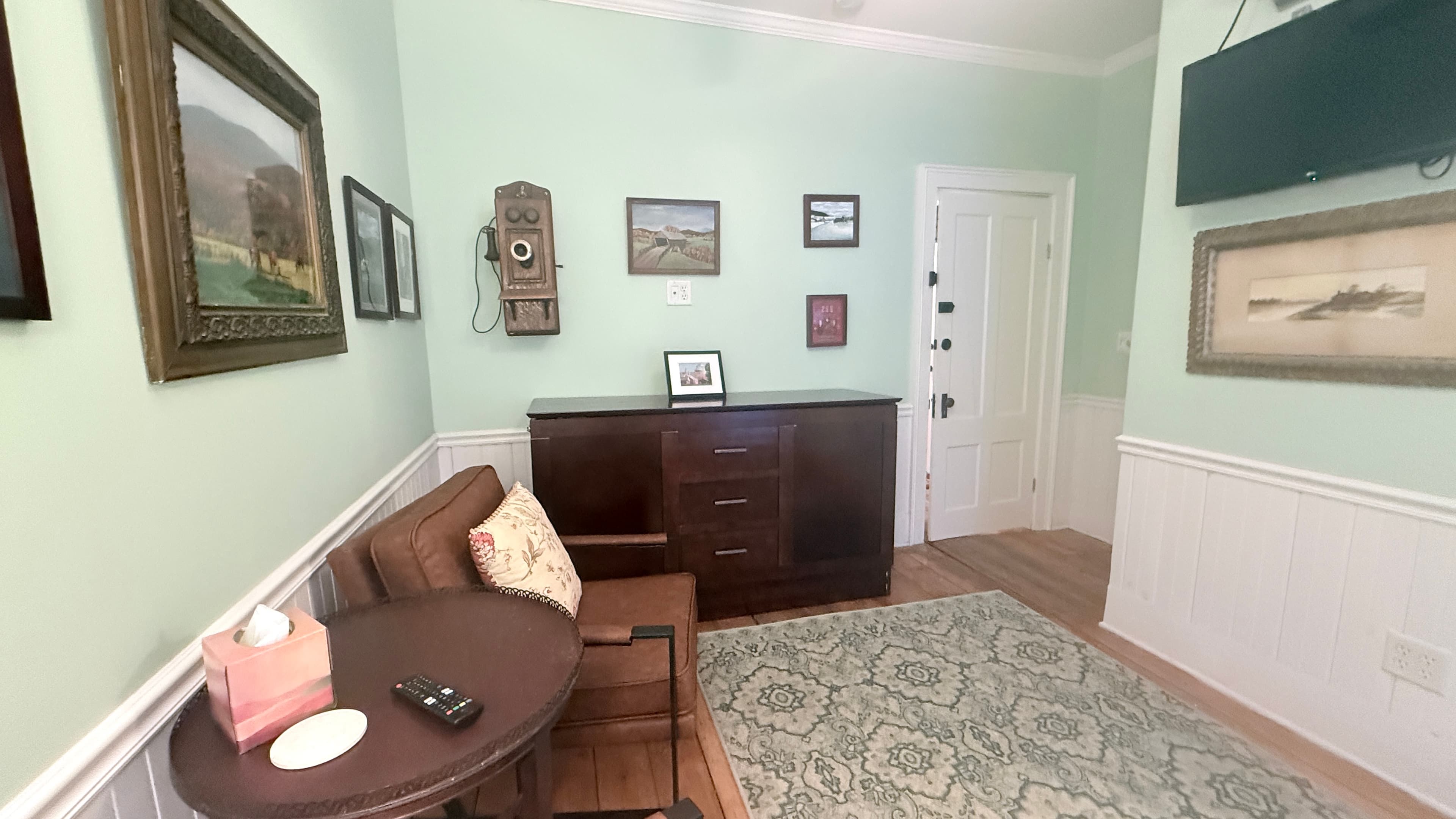 A cozy bedroom area with light green walls and white wainscoting. The corner features two brown armchairs with patterned pillows arranged around a small dark wood table, set on a green and cream patterned area rug. A collection of framed landscape art and a vintage-style wooden wall phone decorate the walls, and a large dark wood dresser is positioned to the right.