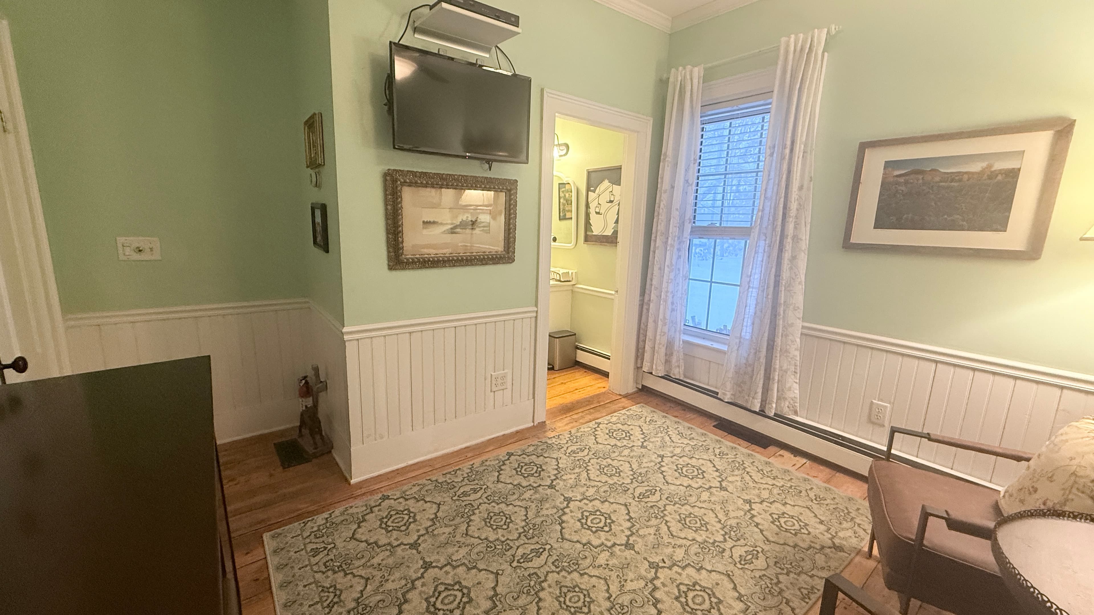 A bright corner of a room with pale green walls and white wainscoting, featuring a large window with sheer white curtains. A television is mounted on the wall above a framed landscape picture, and an open doorway reveals a glimpse into a bathroom. A patterned area rug is visible on the floor next to a dark armchair in the corner.