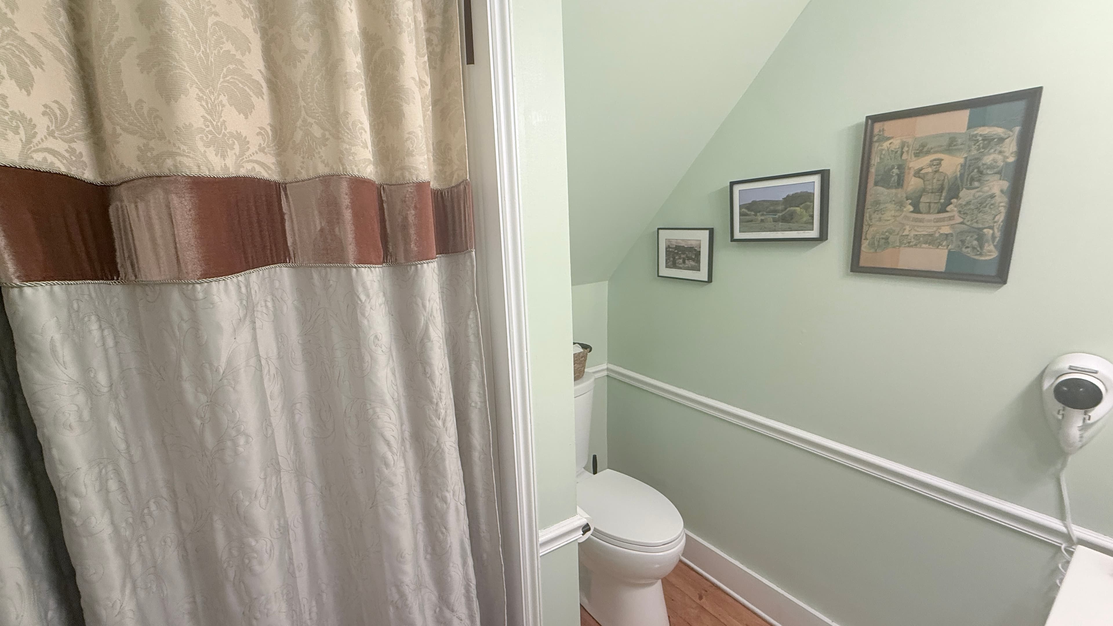 A close-up view of a bathroom corner under a slanted ceiling, featuring light green walls with white wainscoting. A white toilet is tucked into the corner next to a shower area partially obscured by a tan and brown patterned curtain. The wall is decorated with three small framed landscape pictures, and a white hair dryer is mounted on the right.