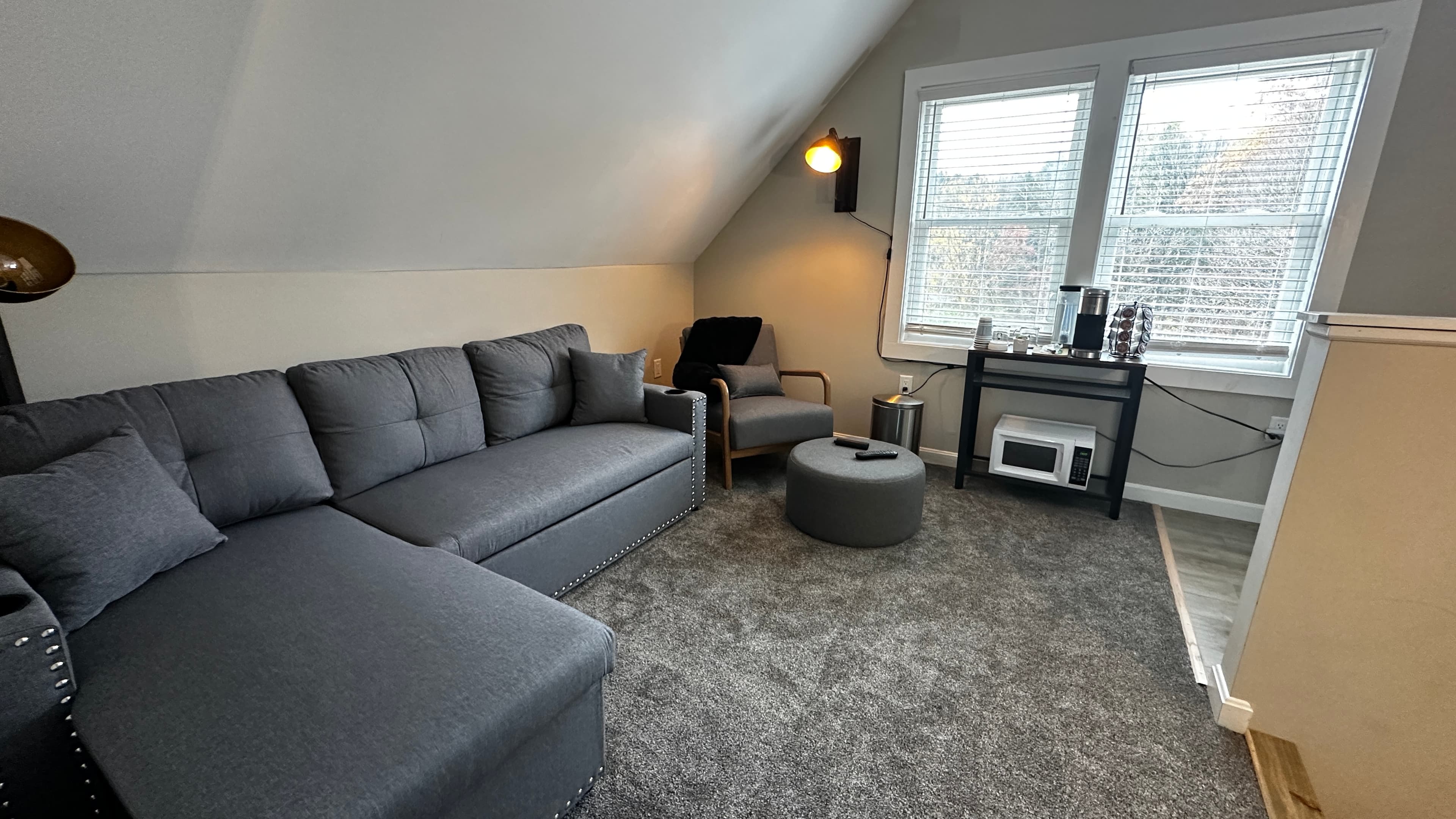 A contemporary living area with a sloped white ceiling and grey carpeting. The room features a large charcoal grey sectional sofa, a matching armchair with a black throw, and a circular ottoman. Near two bright windows, a small black table holds a coffee maker and sits above a white microwave.