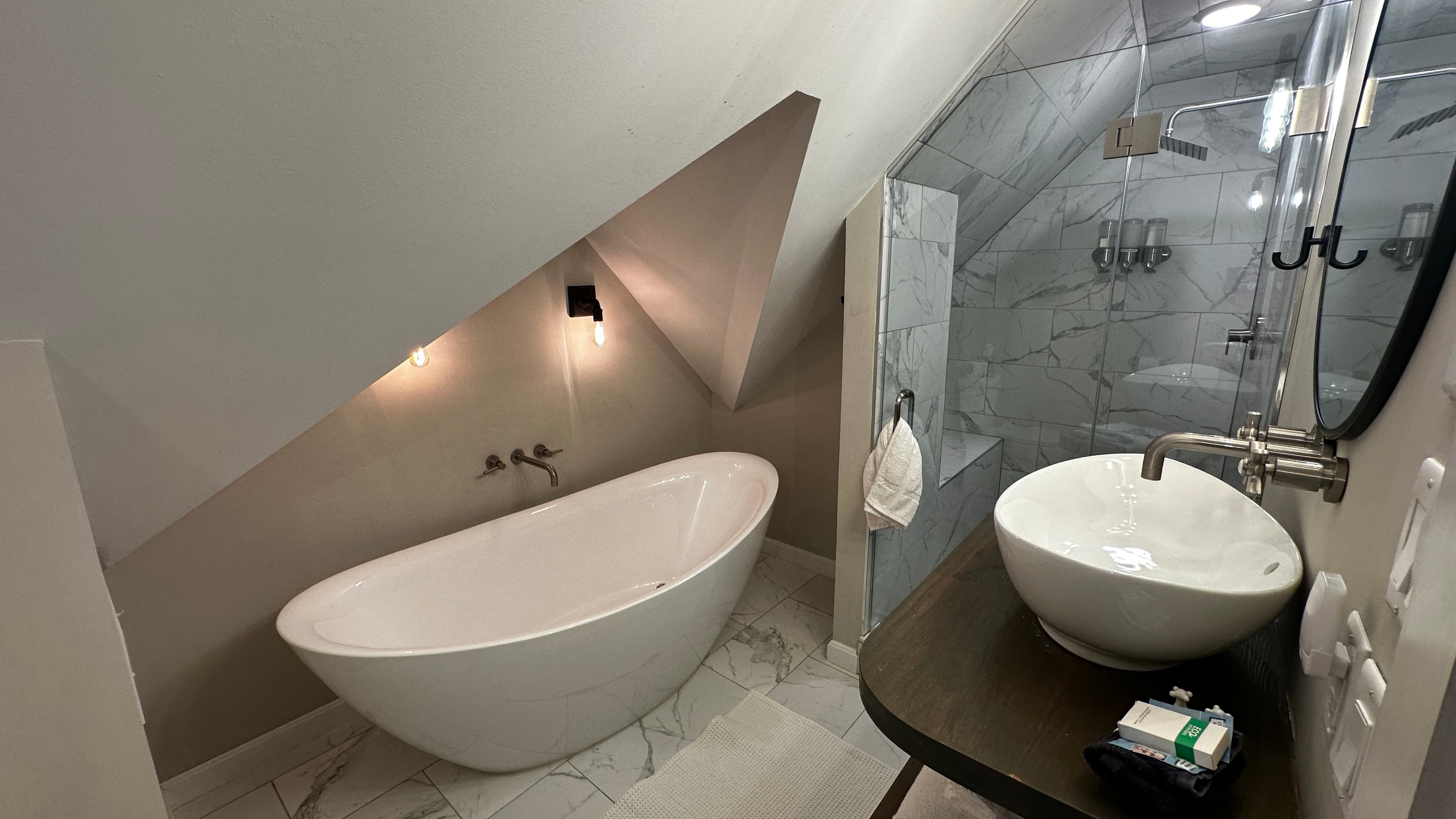 A luxurious, modern bathroom tucked into an attic space with a sharply slanted white ceiling. The room features a large, oval-shaped freestanding soaking tub and a sleek vessel sink on a dark wood vanity. To the right, a glass-enclosed walk-in shower with marble-patterned tile is visible.