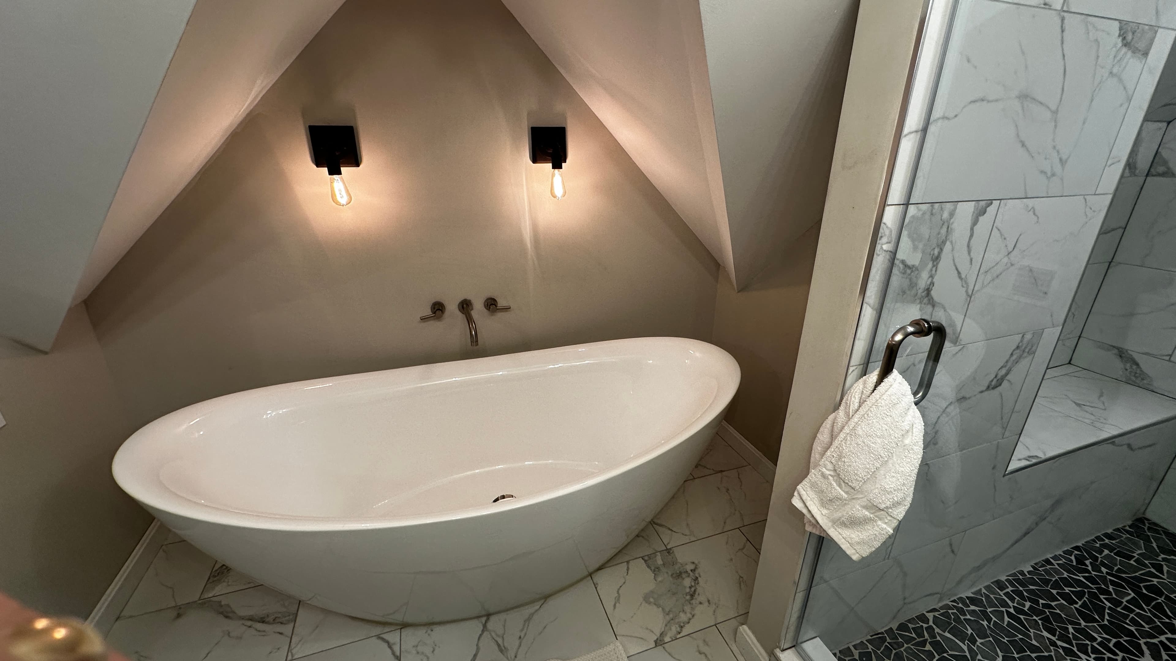 A spacious living area in an attic suite featuring a dark grey sectional sofa and a matching armchair on a plush grey carpet. To the right, a small black utility table sits under two bright windows, and the room is illuminated by warm, modern wall lamps against a slanted white ceiling.